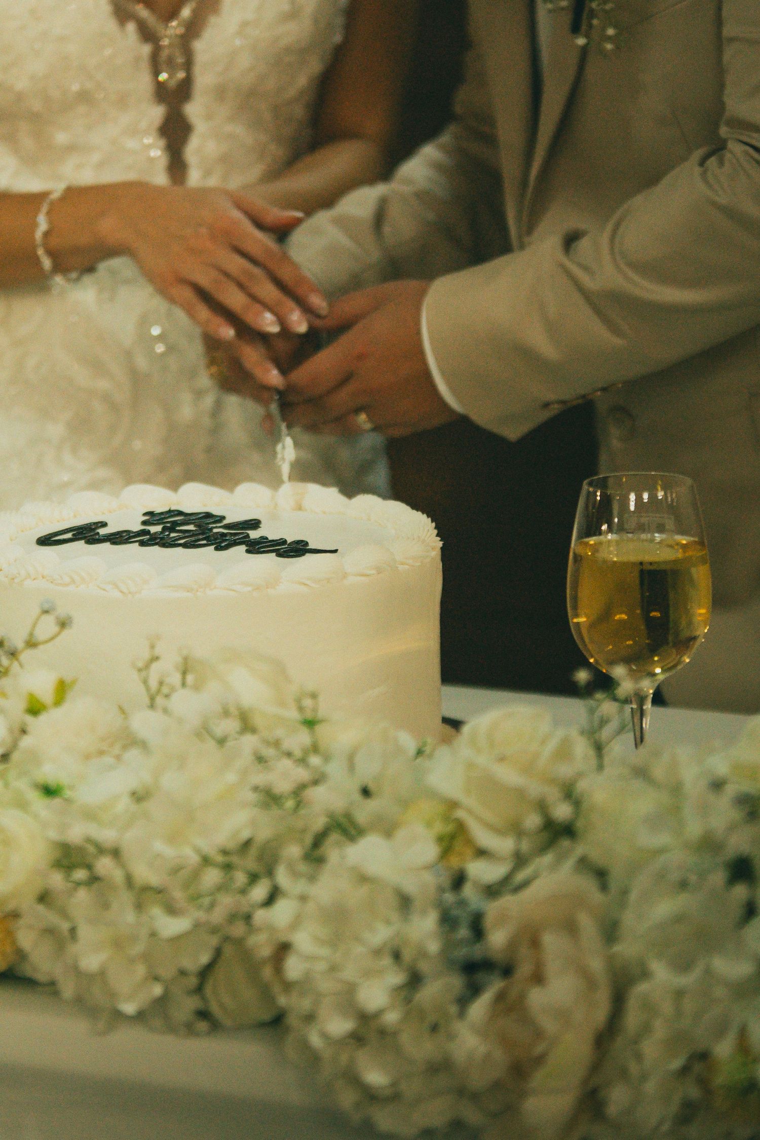 Close-up of a newly married couples' hands cutting white wedding cake decorated with fresh flowers.