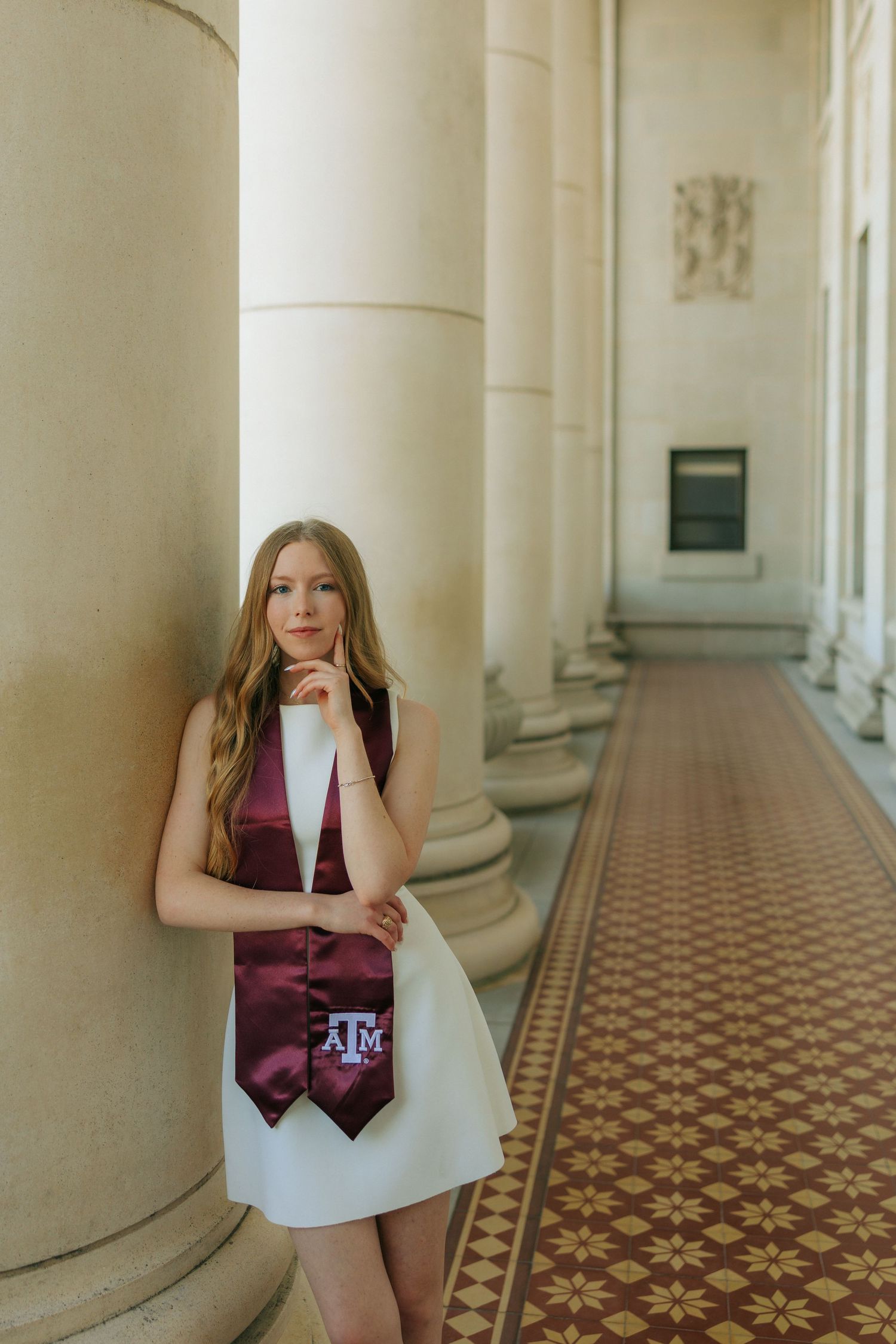 A gradate poses in a white dress in a classical columned hallway.