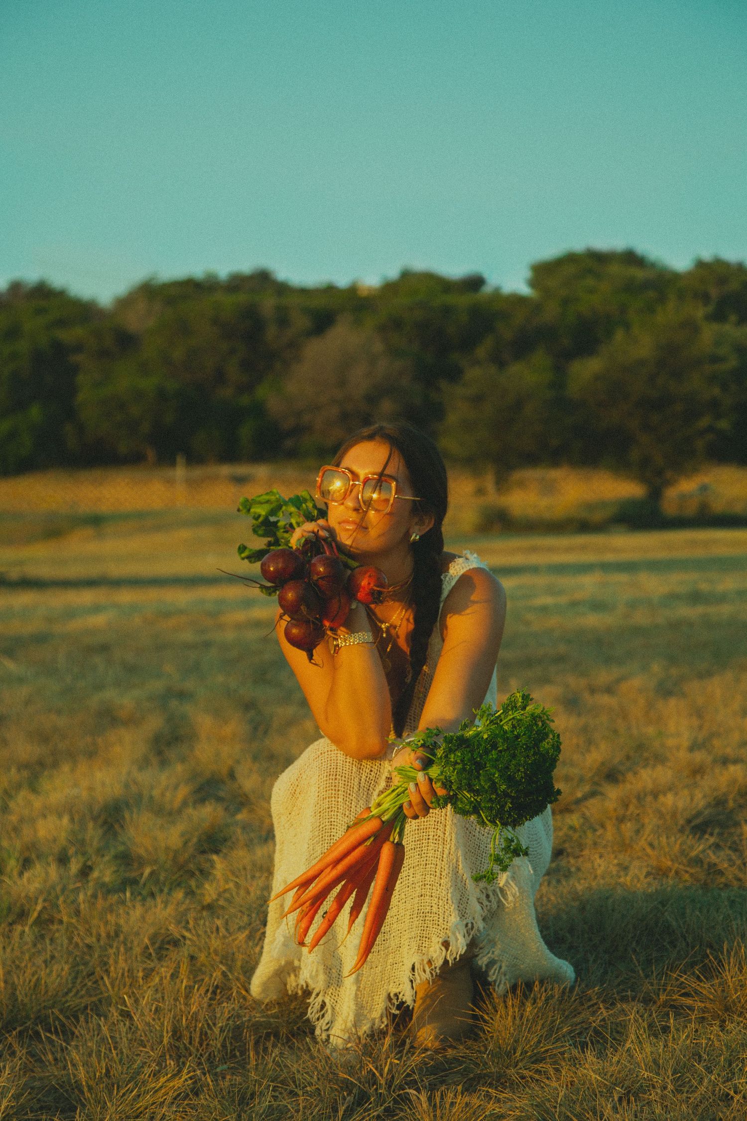 Woman in white dress holds garden-fresh carrots and greens in a playful pose against a turquoise sky.