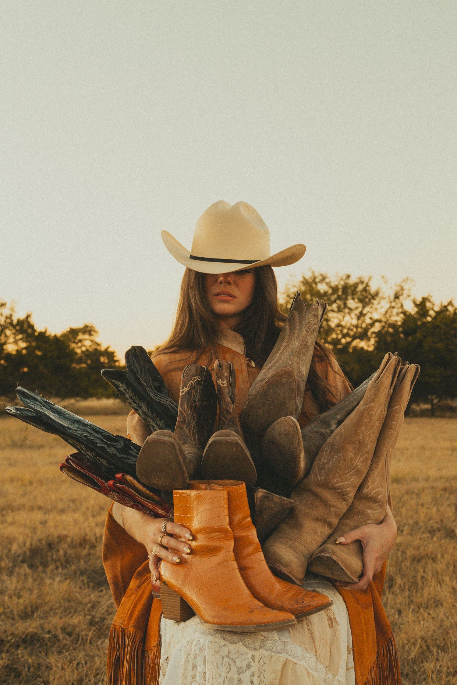 Woman in cowboy hat poses with vintage western boots artistically arranged in golden field.
