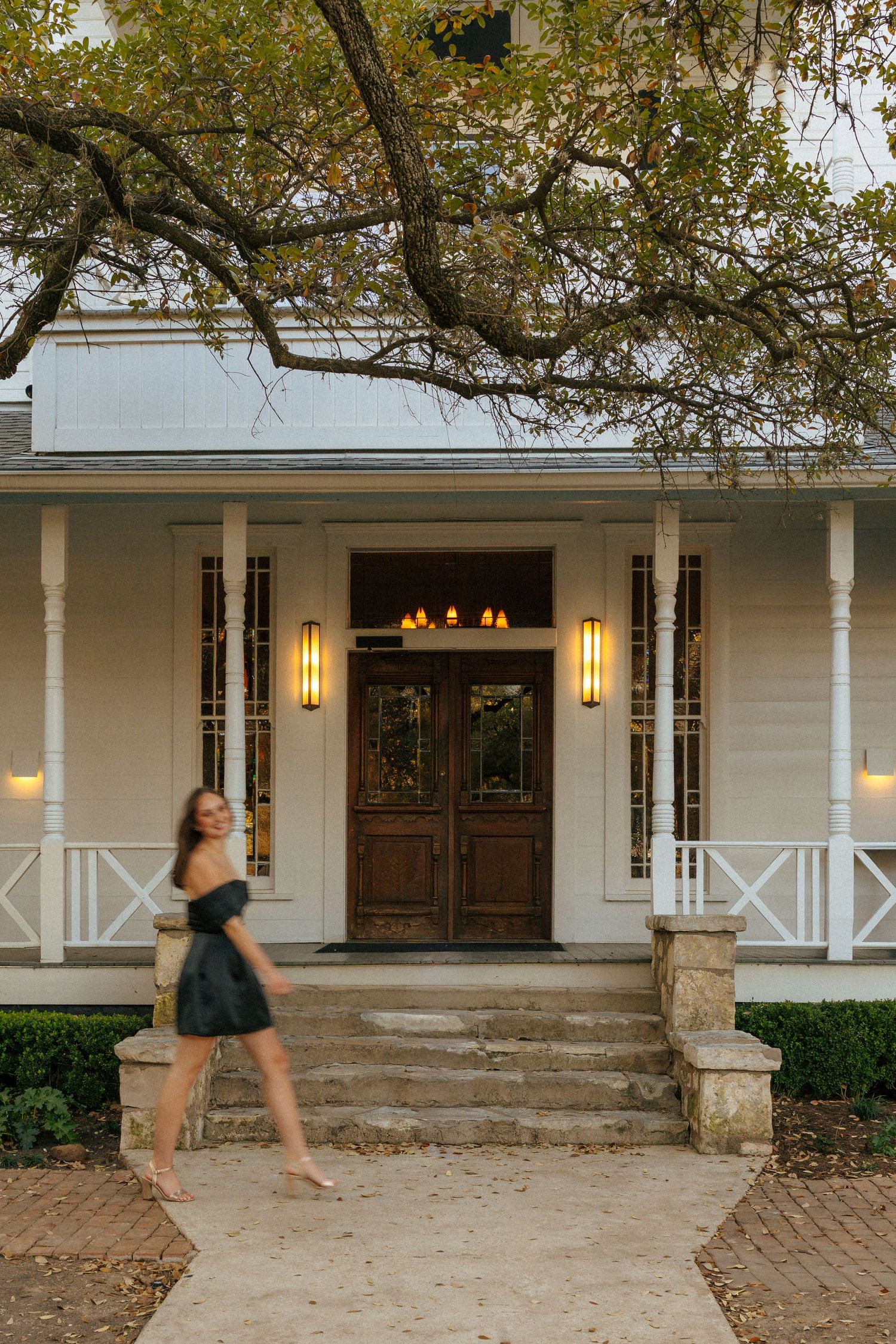 Woman walking in front of white farmhouse with wraparound porch and warm lighting at dusk.