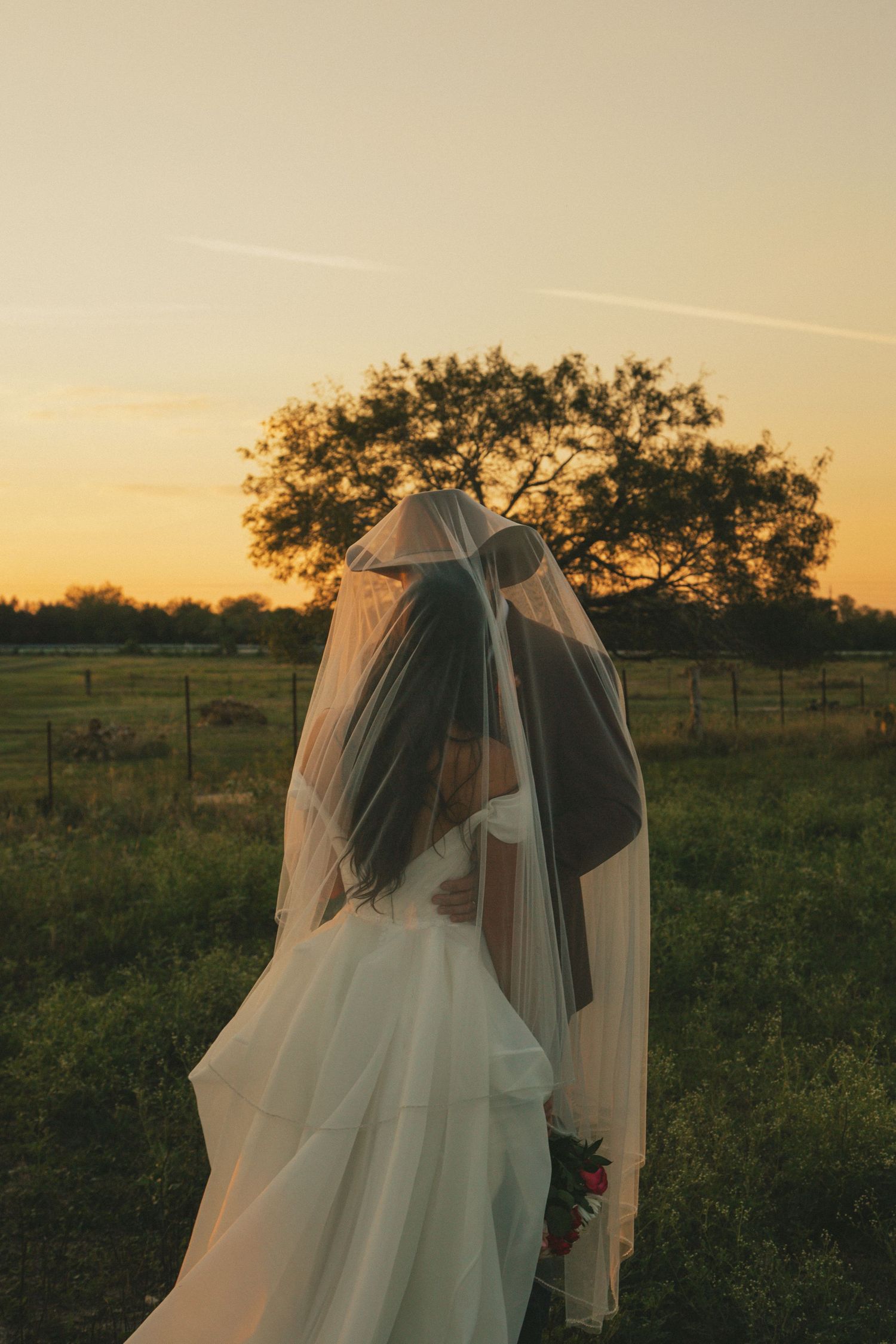 A romantic backlit wedding photo showing a couple embracing under a flowing veil at sunset.