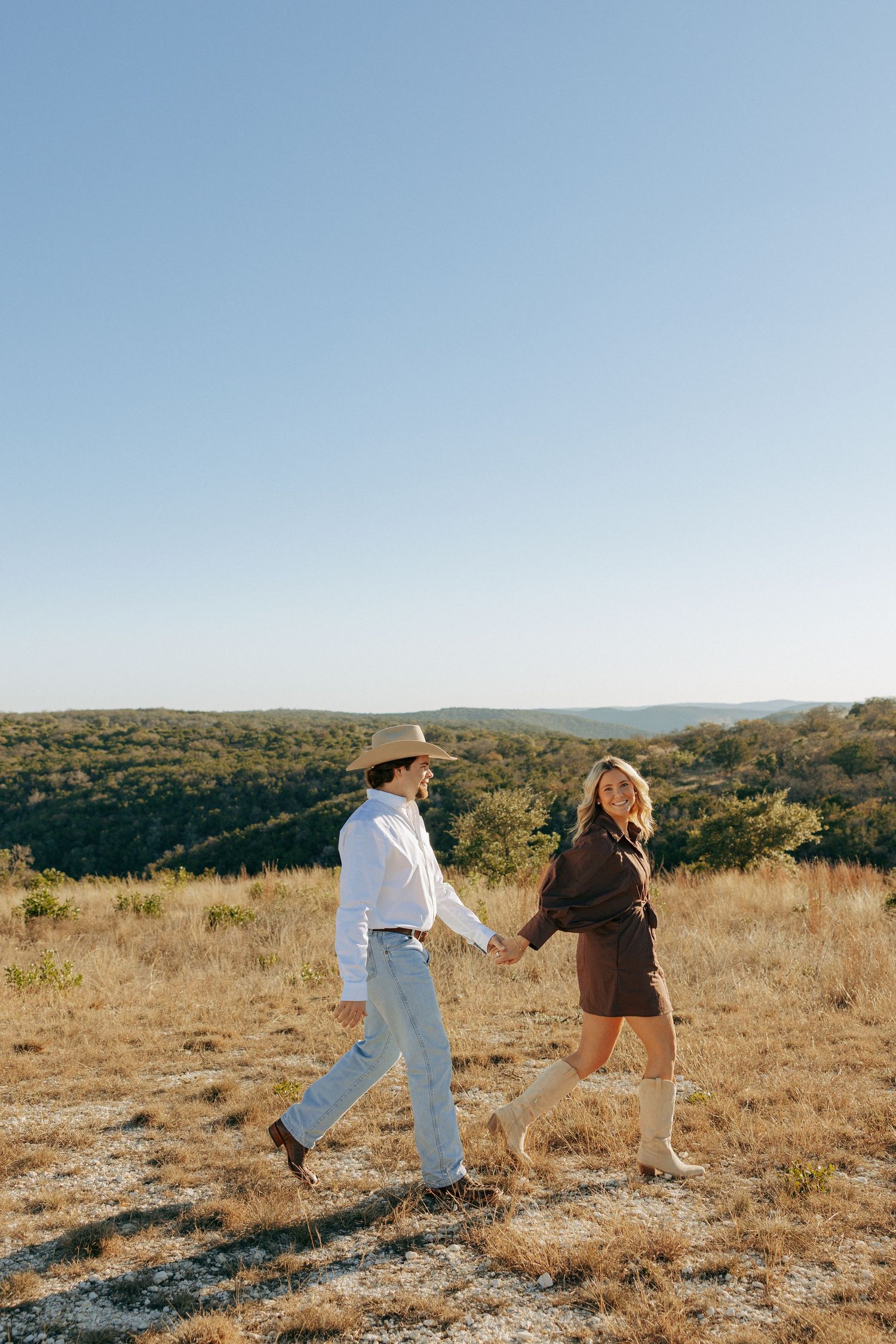 Couple runs playfully through a dry grass field with mountains in the background.