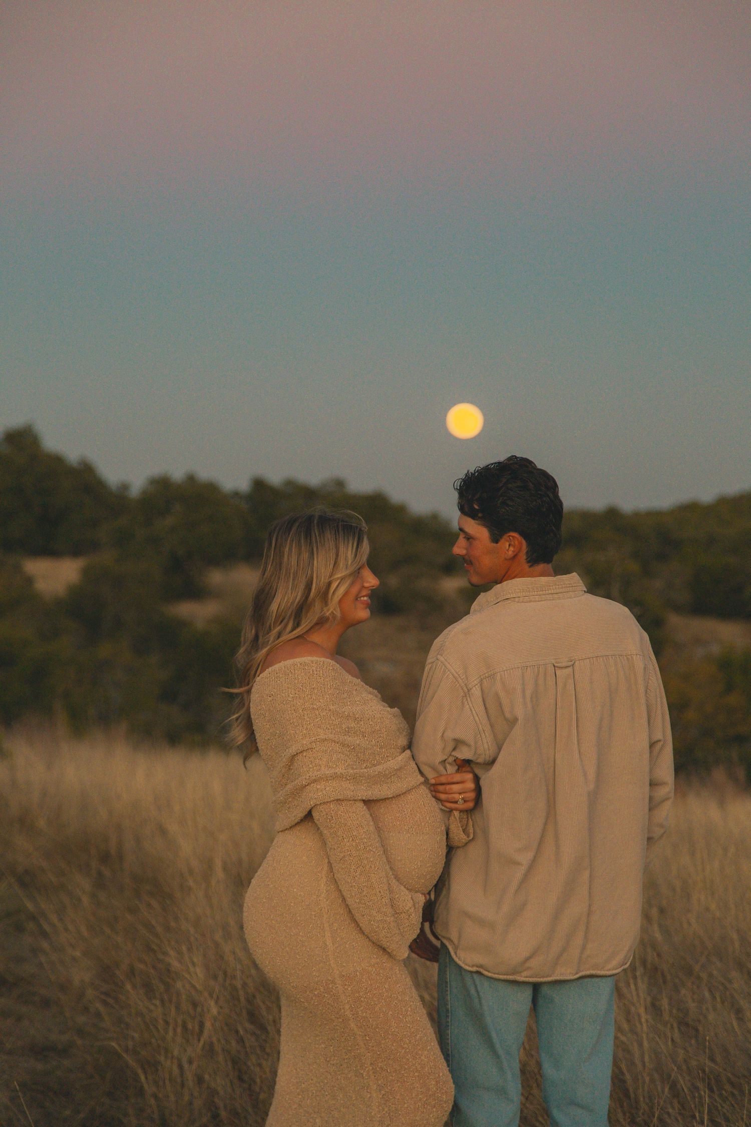Couple standing together in tall grass at sunset with full moon rising.