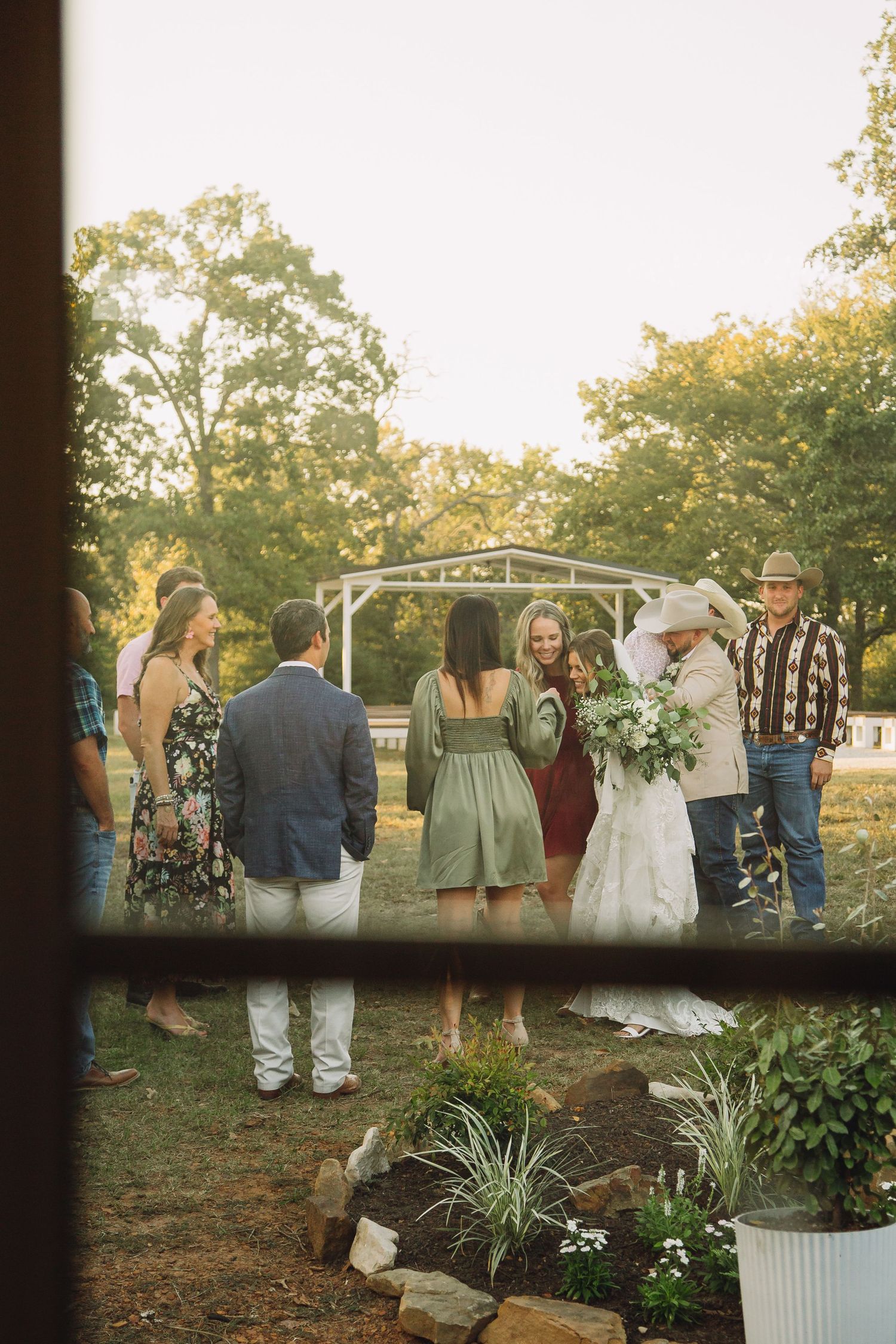 Wedding guests gather outdoors at sunset, viewed through window panes.