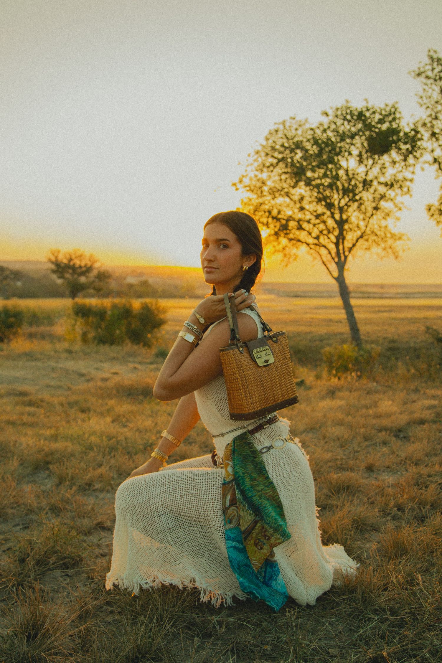 Woman in white dress sitting with basket in rural field at sunset.