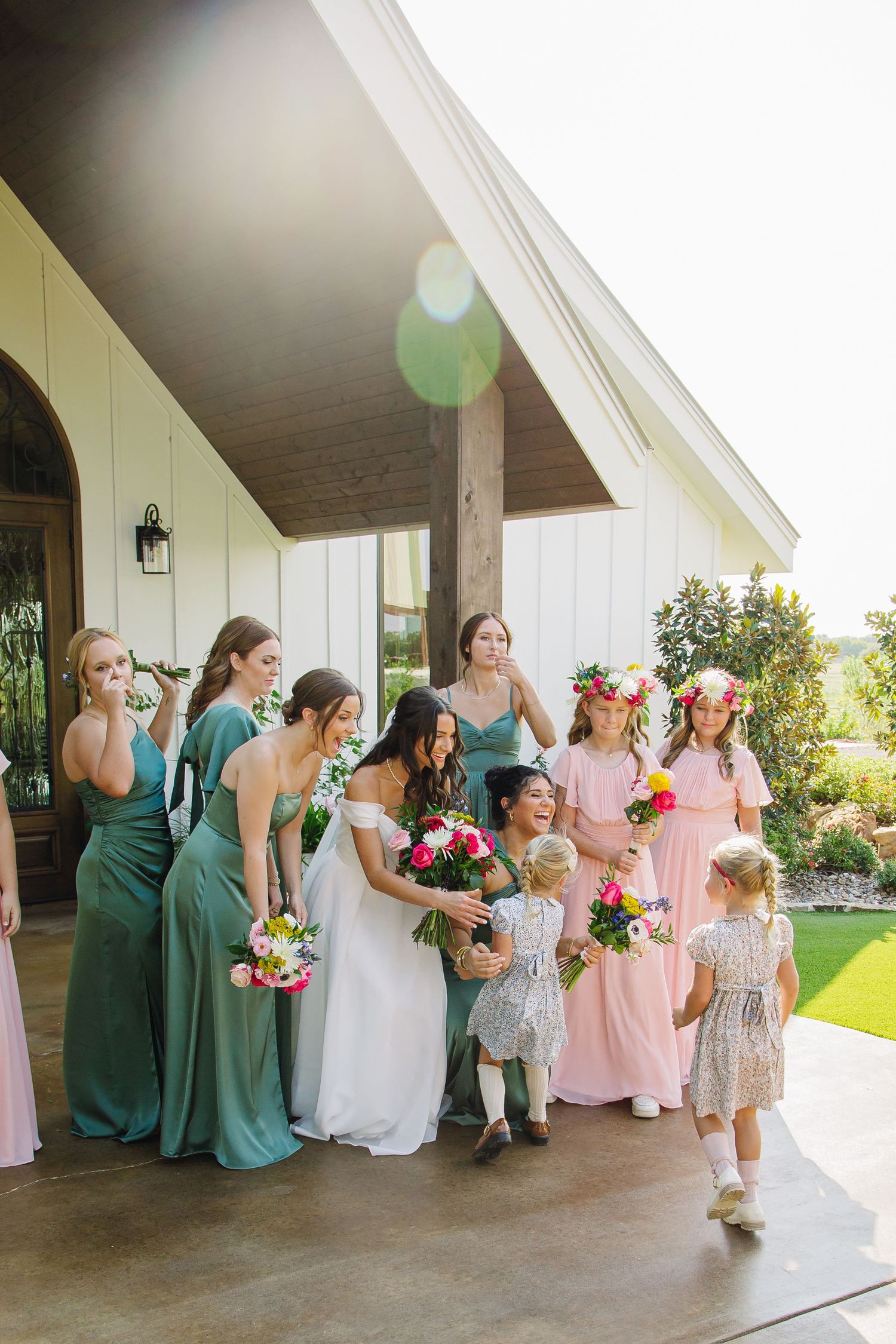 Bridesmaids in mixed pink and green dresses pose together on a white porch.
