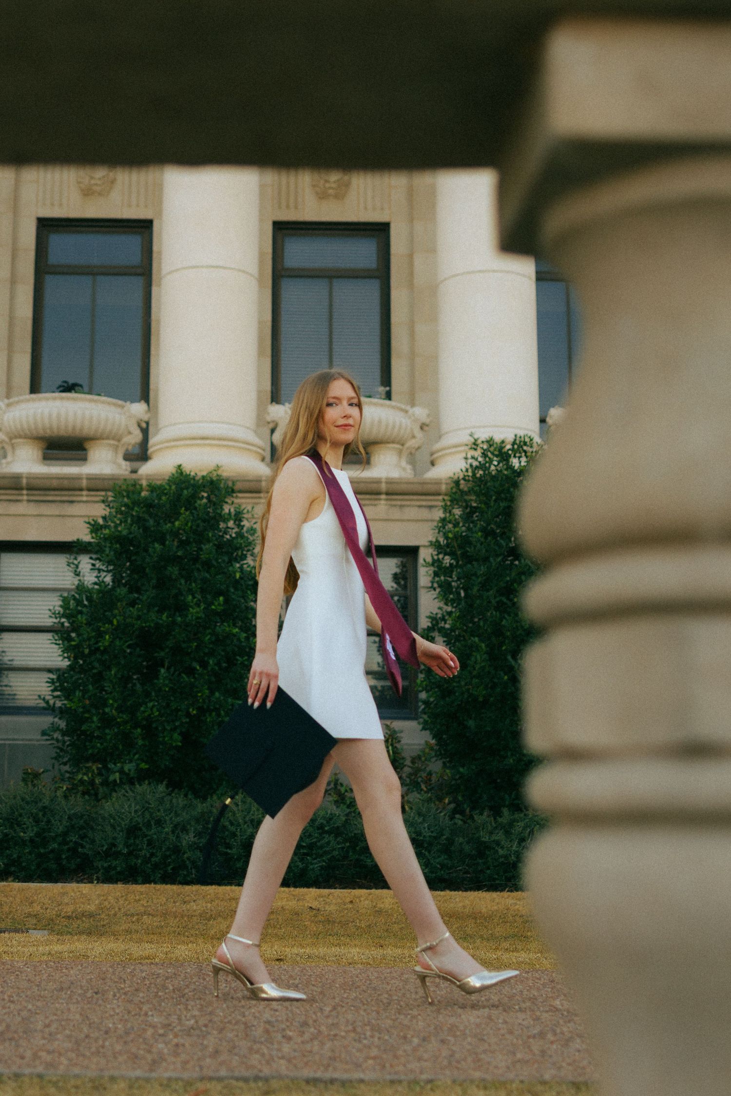 A graduate in a white dress walking past architectural columns on a sunny day.