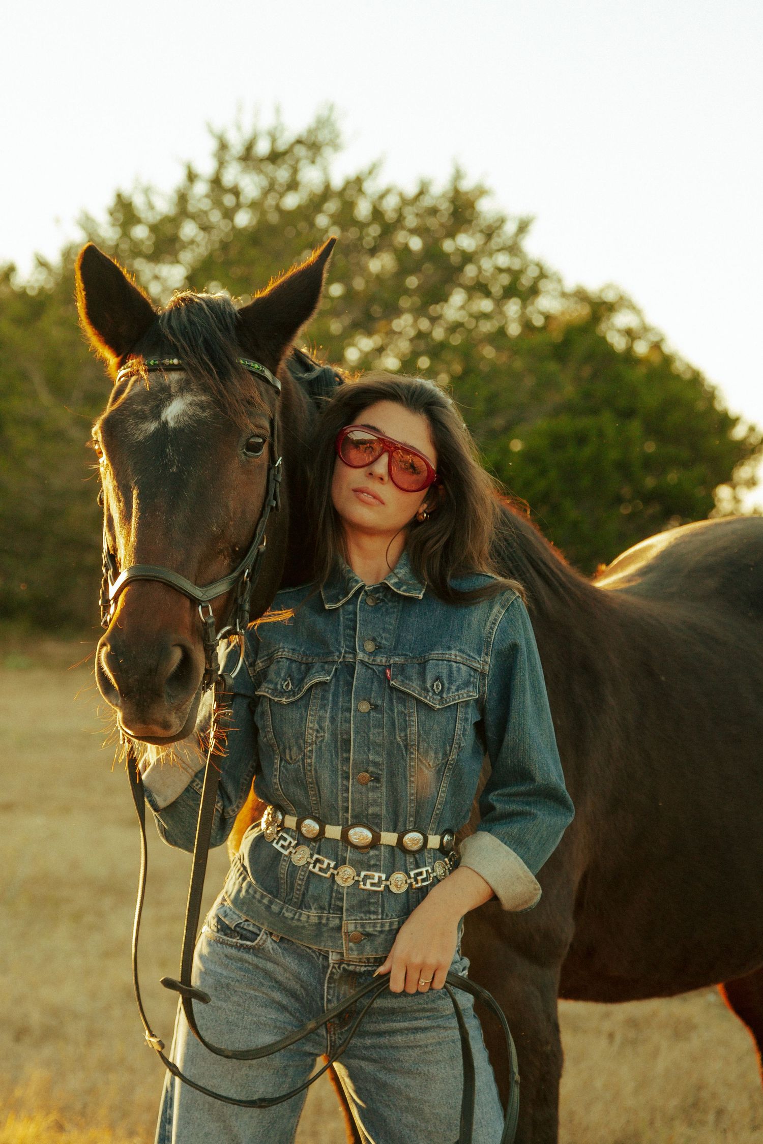Stylish western portrait with a horse featuring a woman in denim outfit, decorative belt, and retro sunglasses.