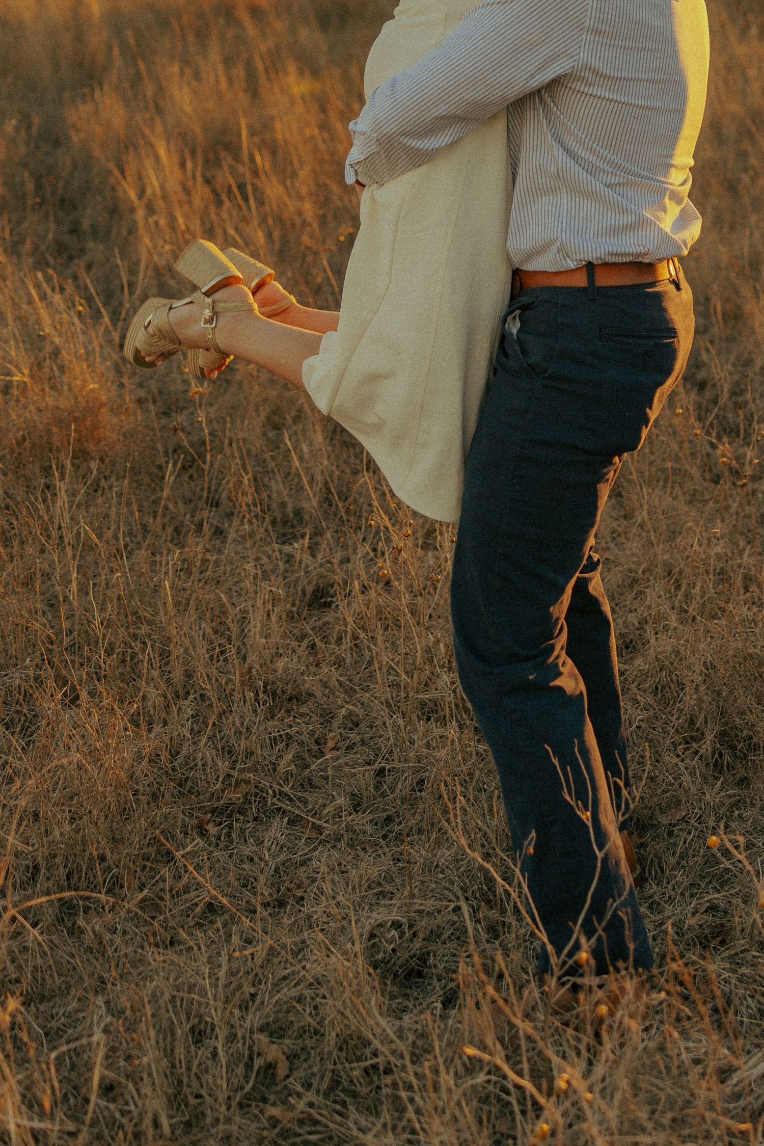 Lower body shot of couple standing close together in tall grass at golden hour.