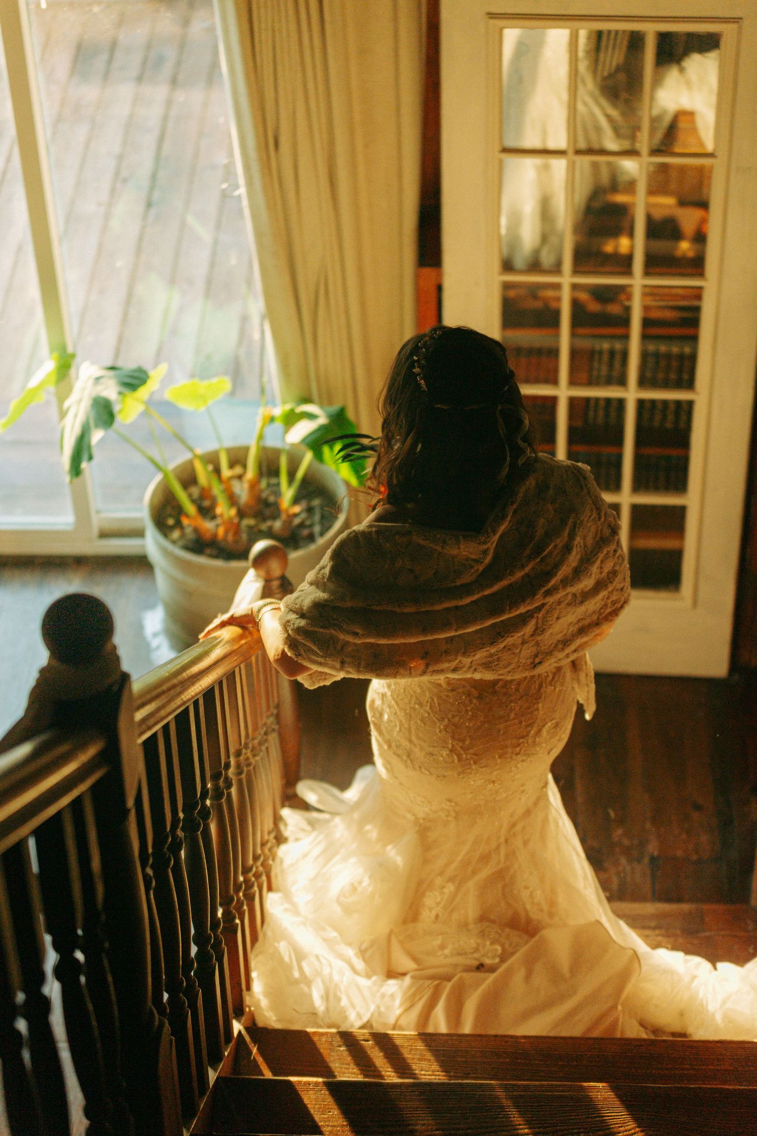 A bride in her wedding dress walks down wooden stairs in warm indoor lighting.