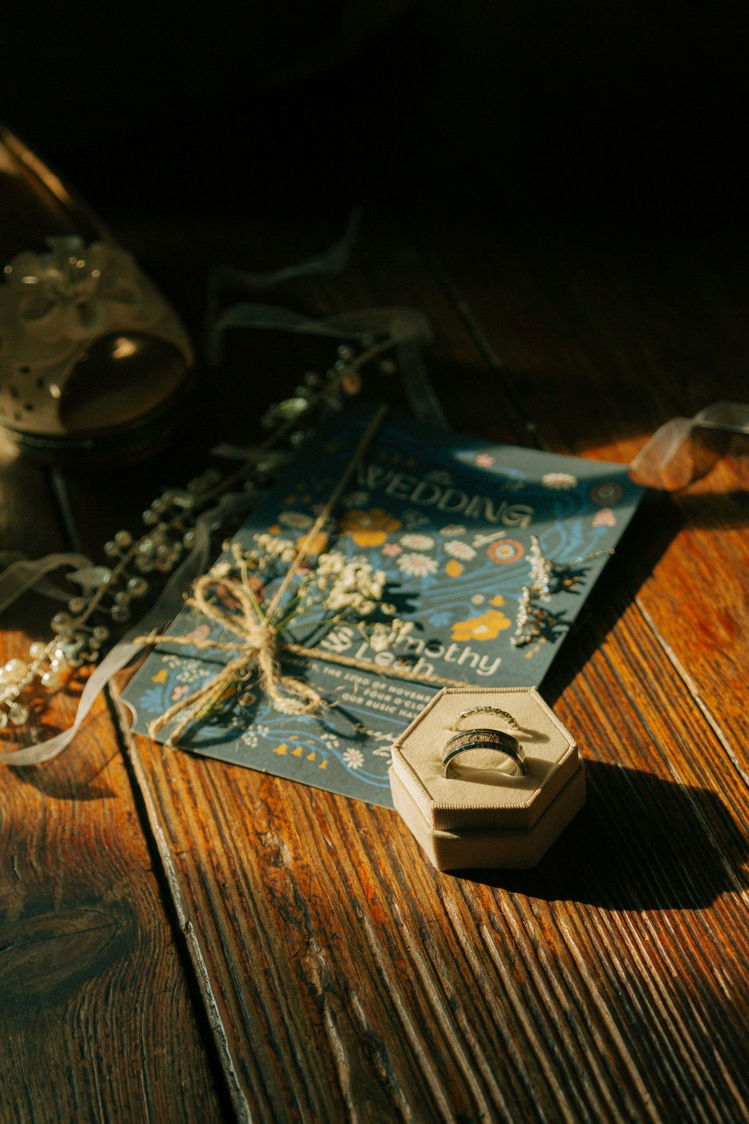 Wedding invitation, wedding bands, and a white elegant high heel arranged artistically on a rustic wooden table in warm lighting.