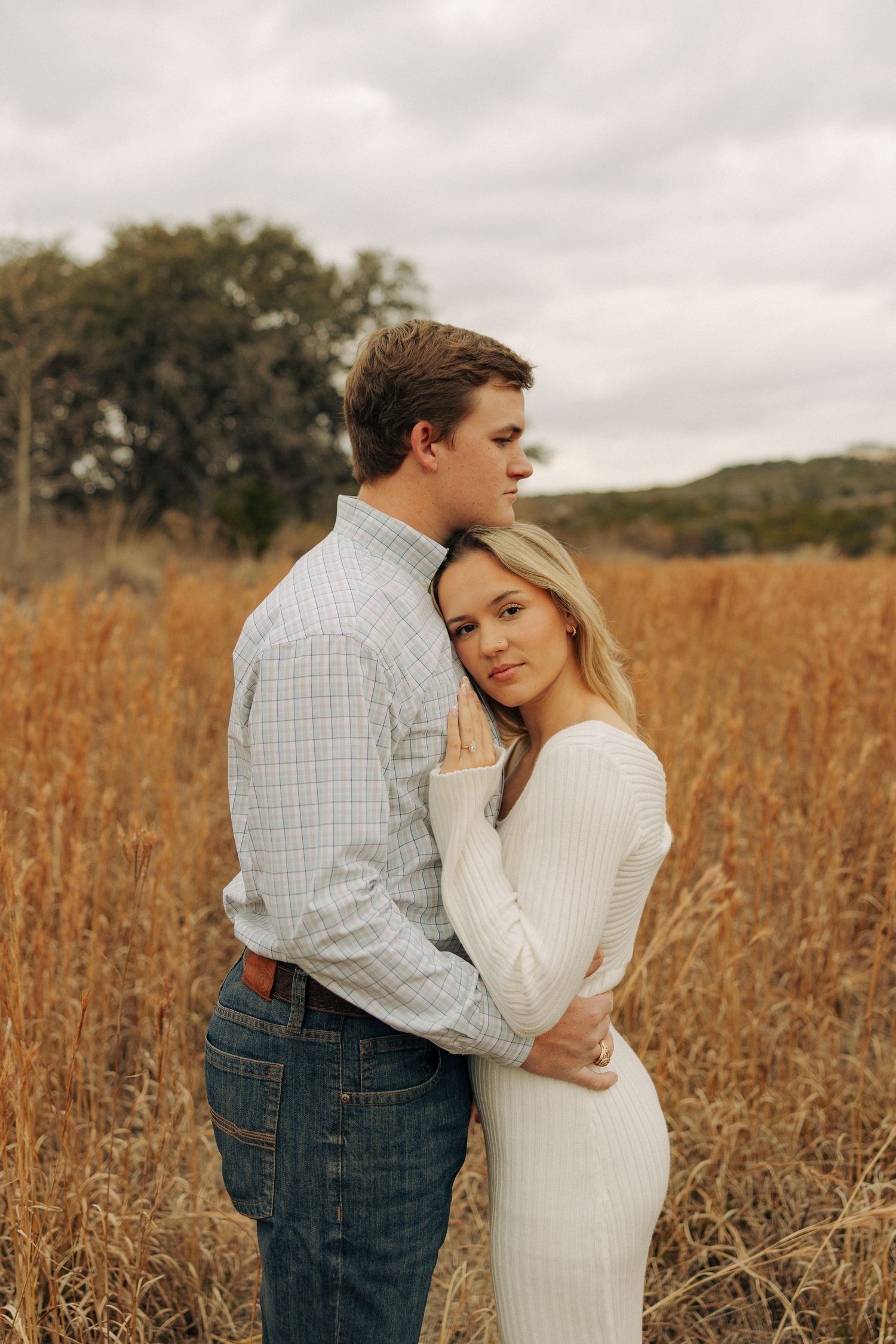 A romantic couple embraces in a golden field during autumn with trees in the background.