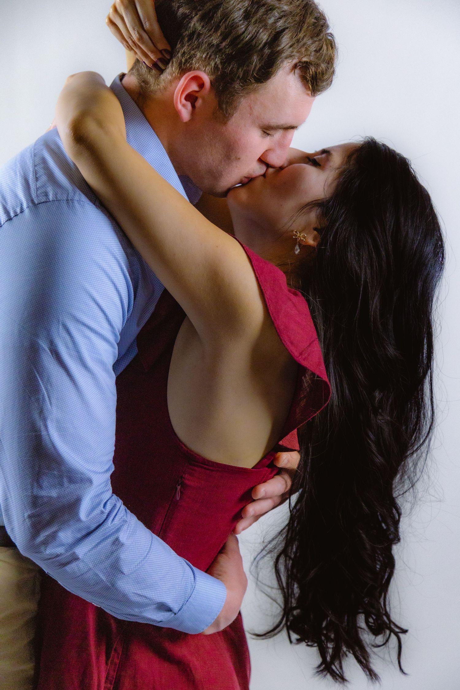 A couple shares a romantic kiss, with the woman in a backless red dress embracing her partner.
