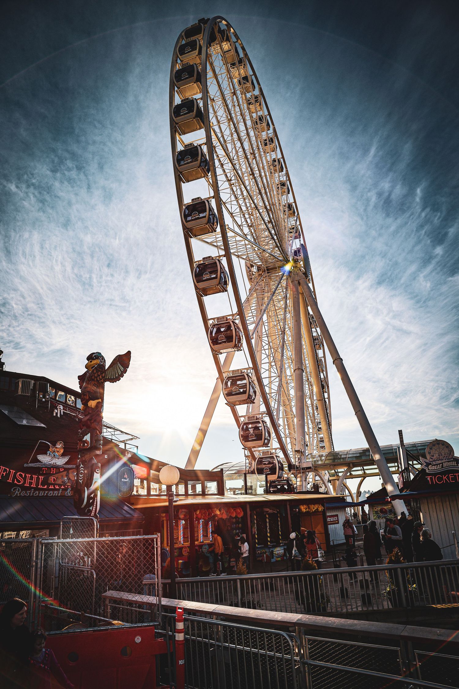A towering Ferris wheel rises above a colorful fairground with carnival stalls and visitors below at dusk.