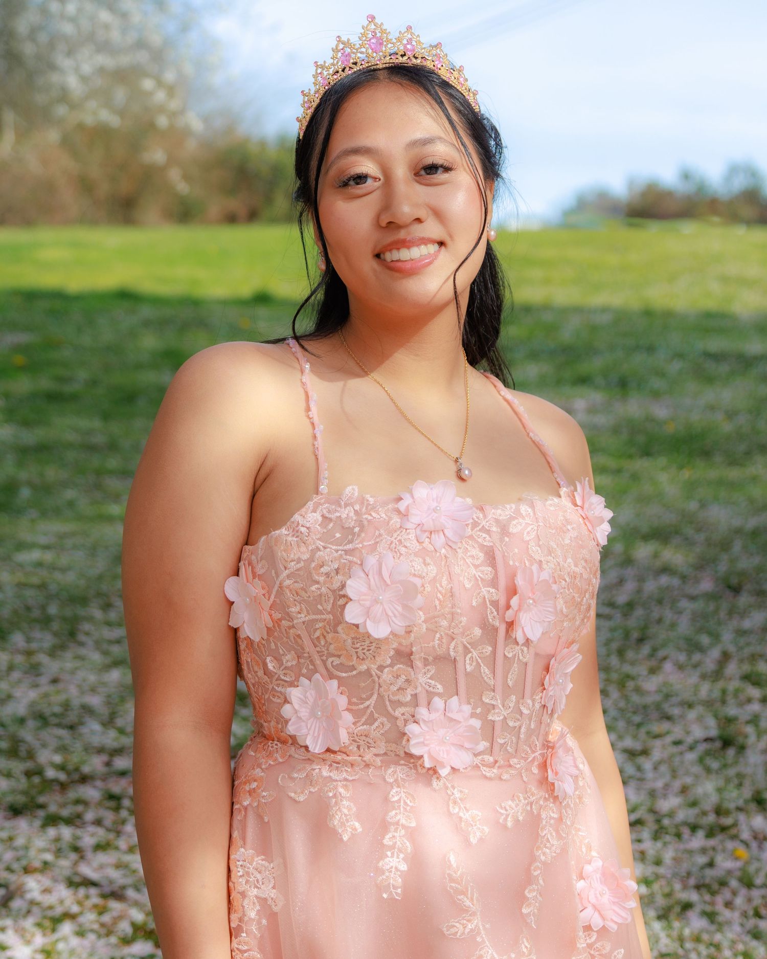 Young woman in pink floral gown and tiara smiling outdoors in a green field on a sunny day.