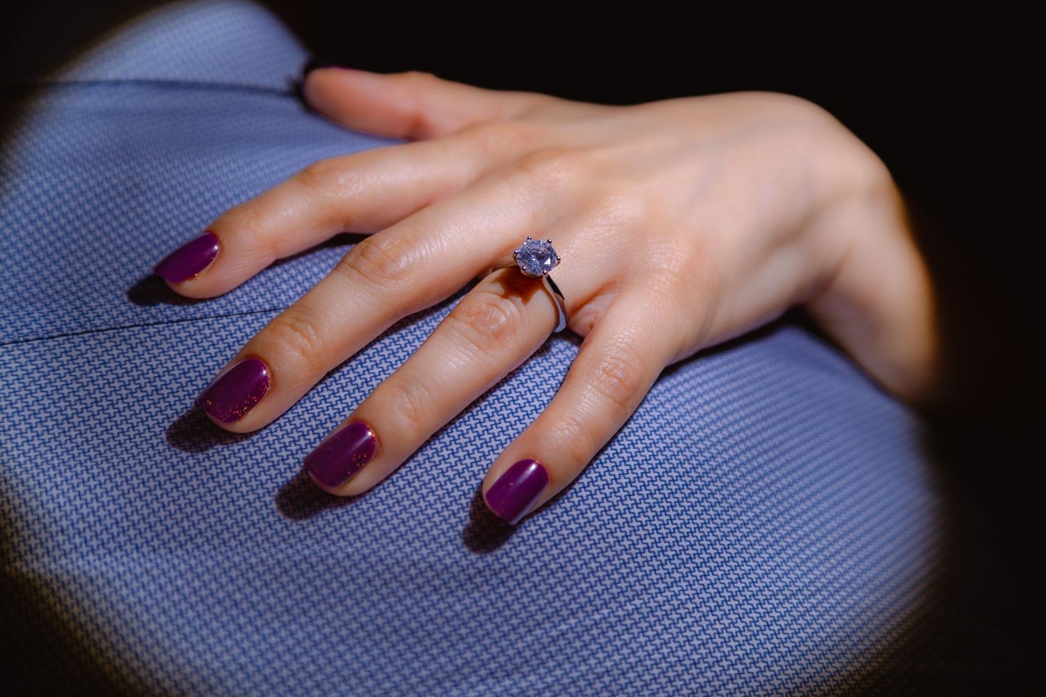 Close-up of a diamond engagement ring on a hand with purple nails resting on blue fabric.