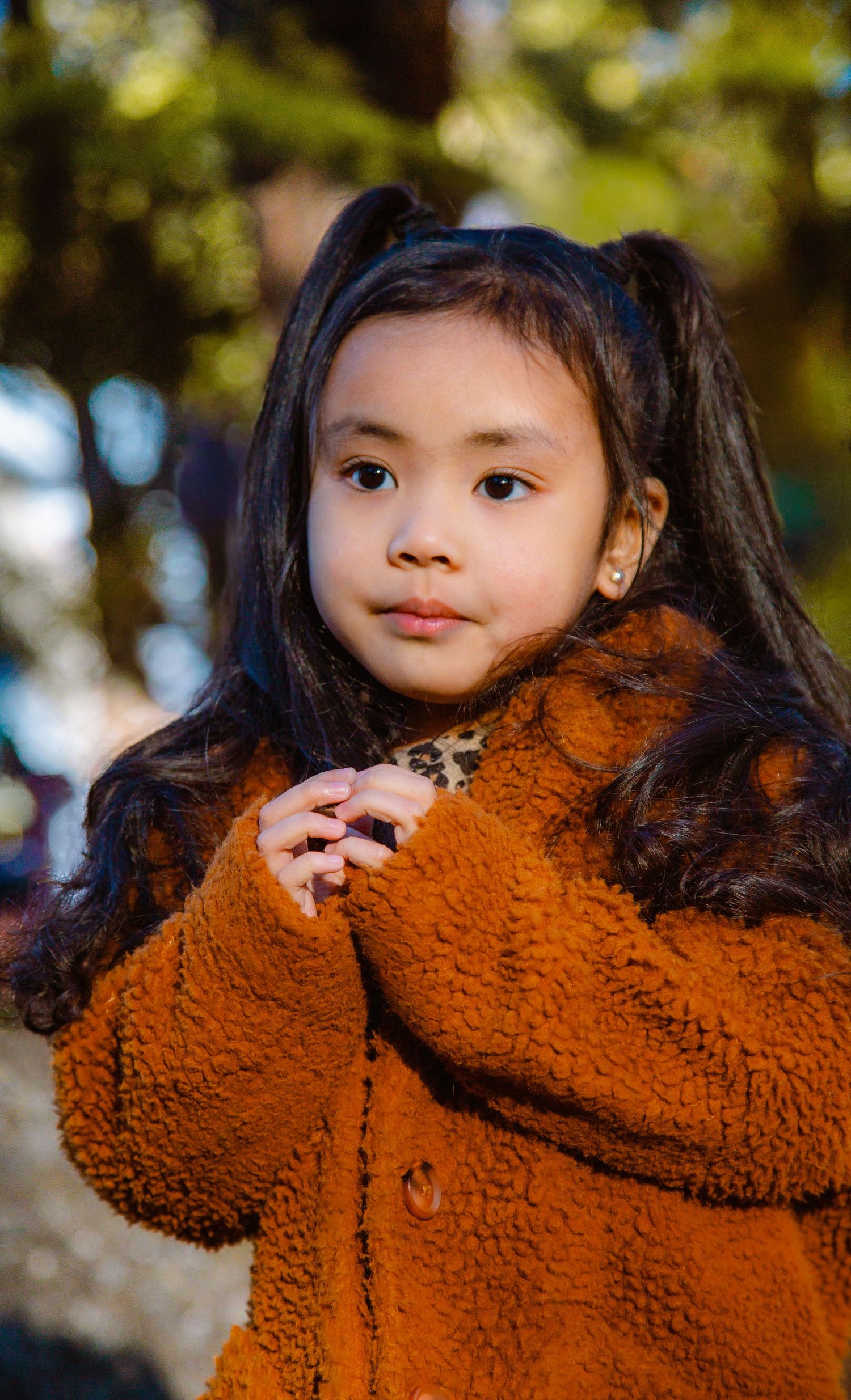 Adorable young Asian girl in orange teddy coat with pigtails posing outdoors in autumn.