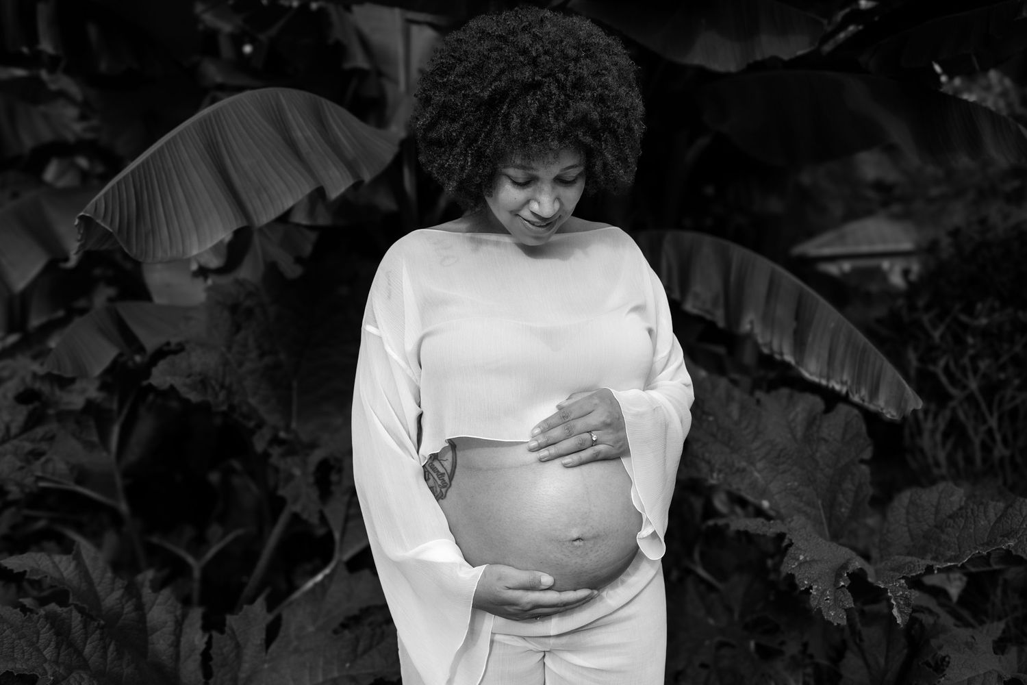 Black and white portrait of pregnant woman in white outfit cradling baby bump among tropical leaves.