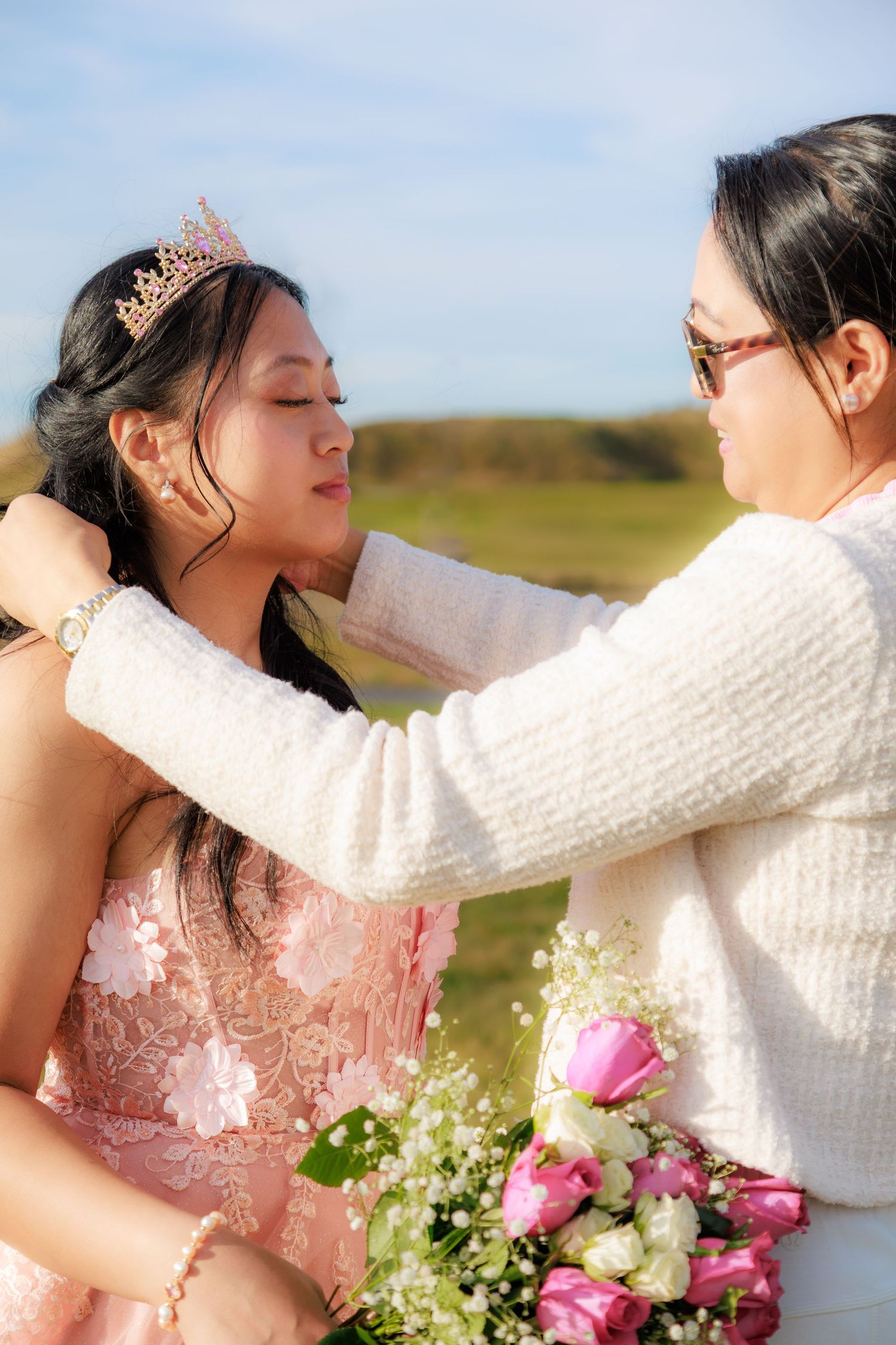 Woman placing a rose gold tiara on a young girl in a pink dress holding a bouquet of pink roses outdoors.