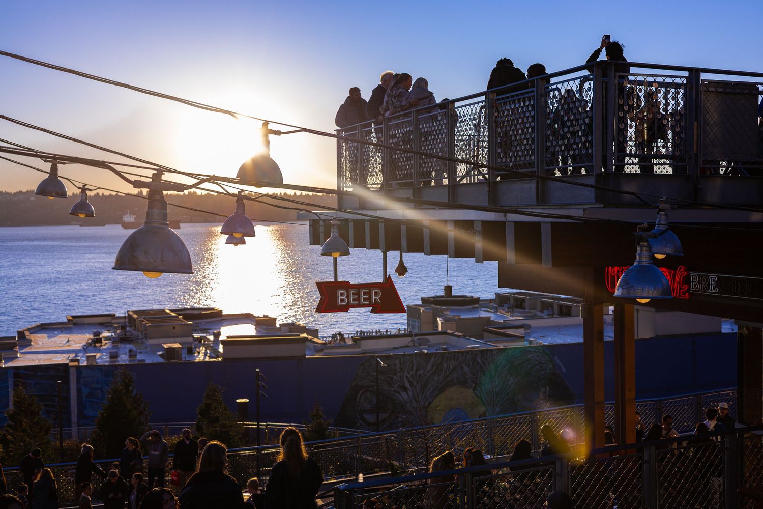 People gather on a waterfront terrace at sunset, with a beer sign and boats visible on the river.