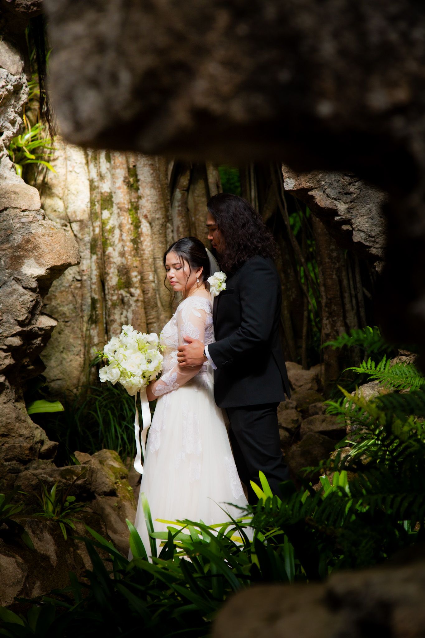 Bride and groom embrace in a lush cave setting, bride holding white bouquet in elegant lace gown.