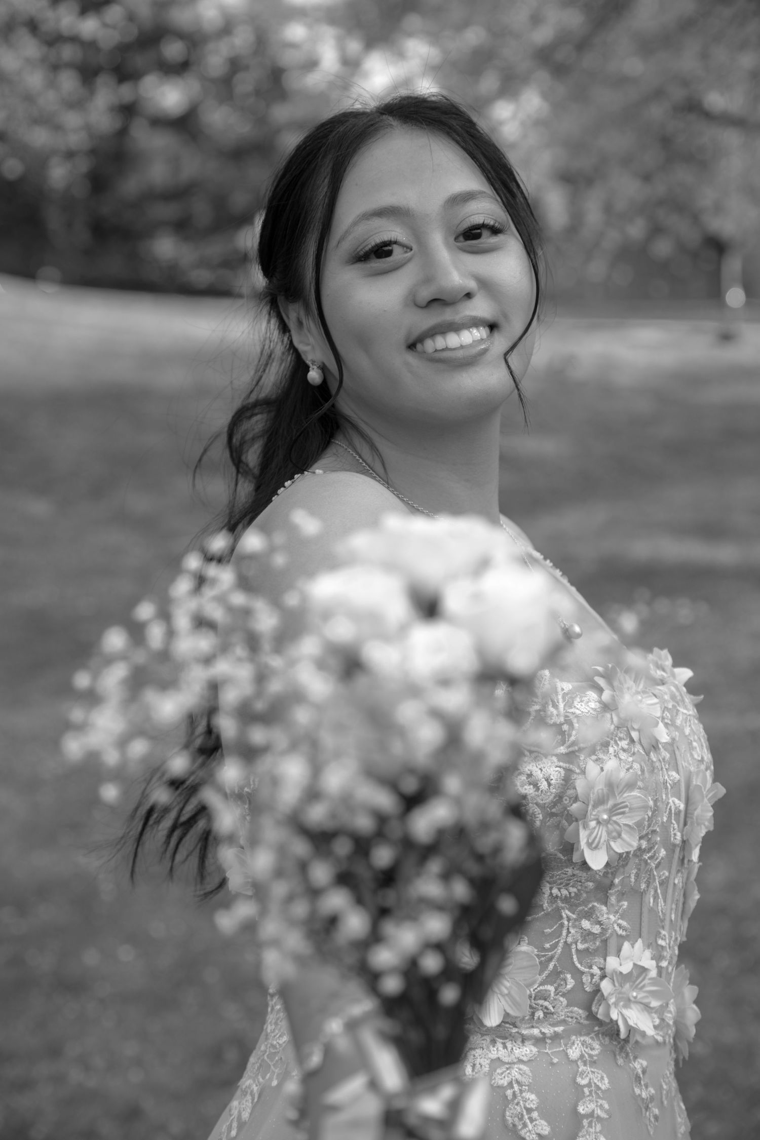 Smiling bride in lace wedding dress holding a bouquet of flowers in a black and white outdoor portrait.