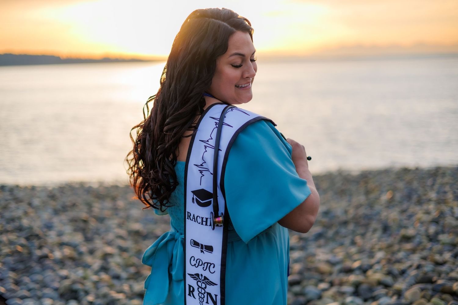 A nursing graduate poses at sunset on a rocky beach wearing a teal gown and BSN graduation sash.