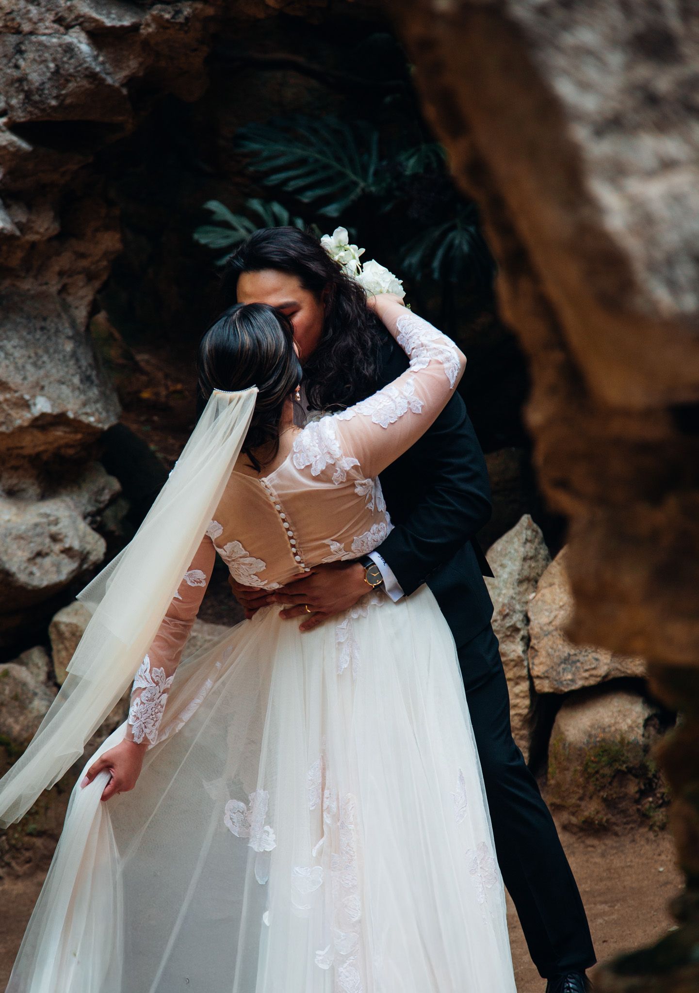 Bride and groom embracing in a romantic cave setting, bride wearing lace wedding gown with veil and floral hair accessories.