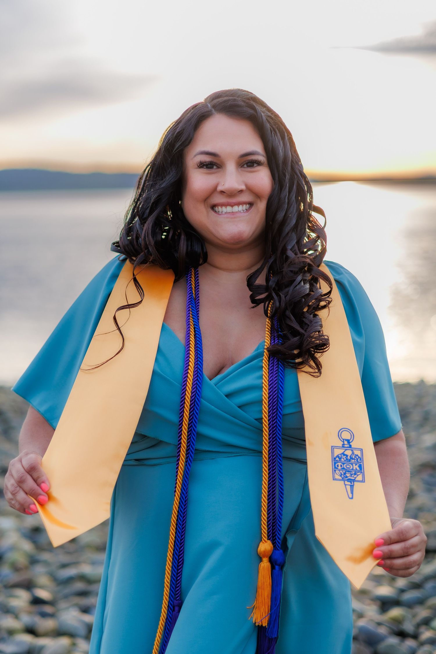 Smiling graduate wearing teal dress with gold honor stole and blue cords poses on rocky beach at sunset.