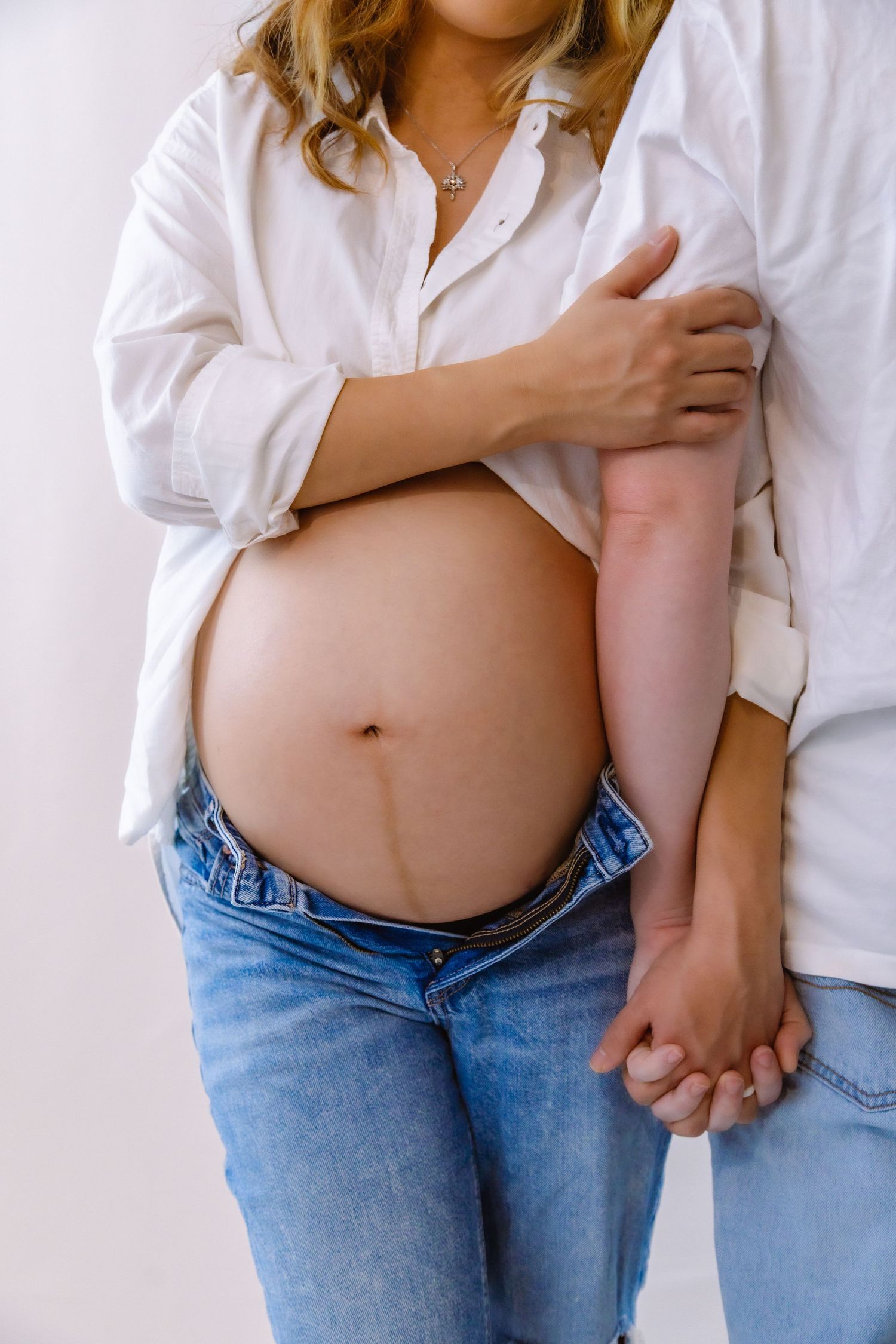 Close-up of pregnant belly with partner holding hands, both wearing white shirts and blue jeans.