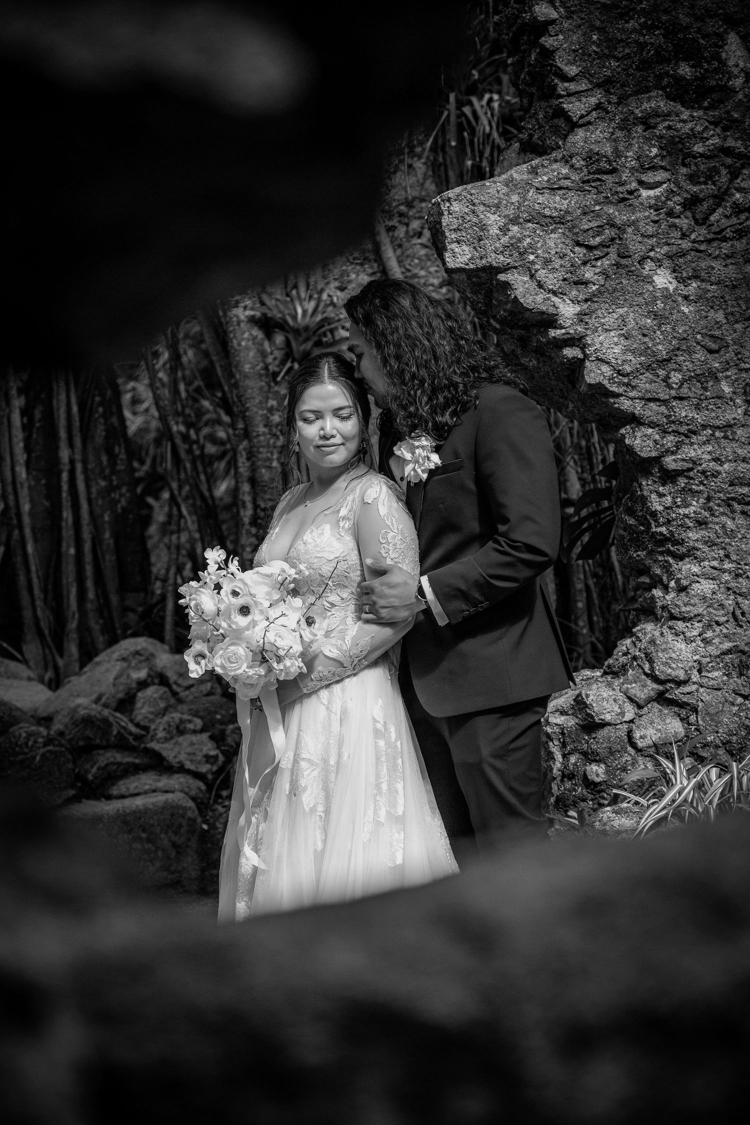 Black and white wedding photo of bride holding bouquet with groom near a rustic stone wall.
