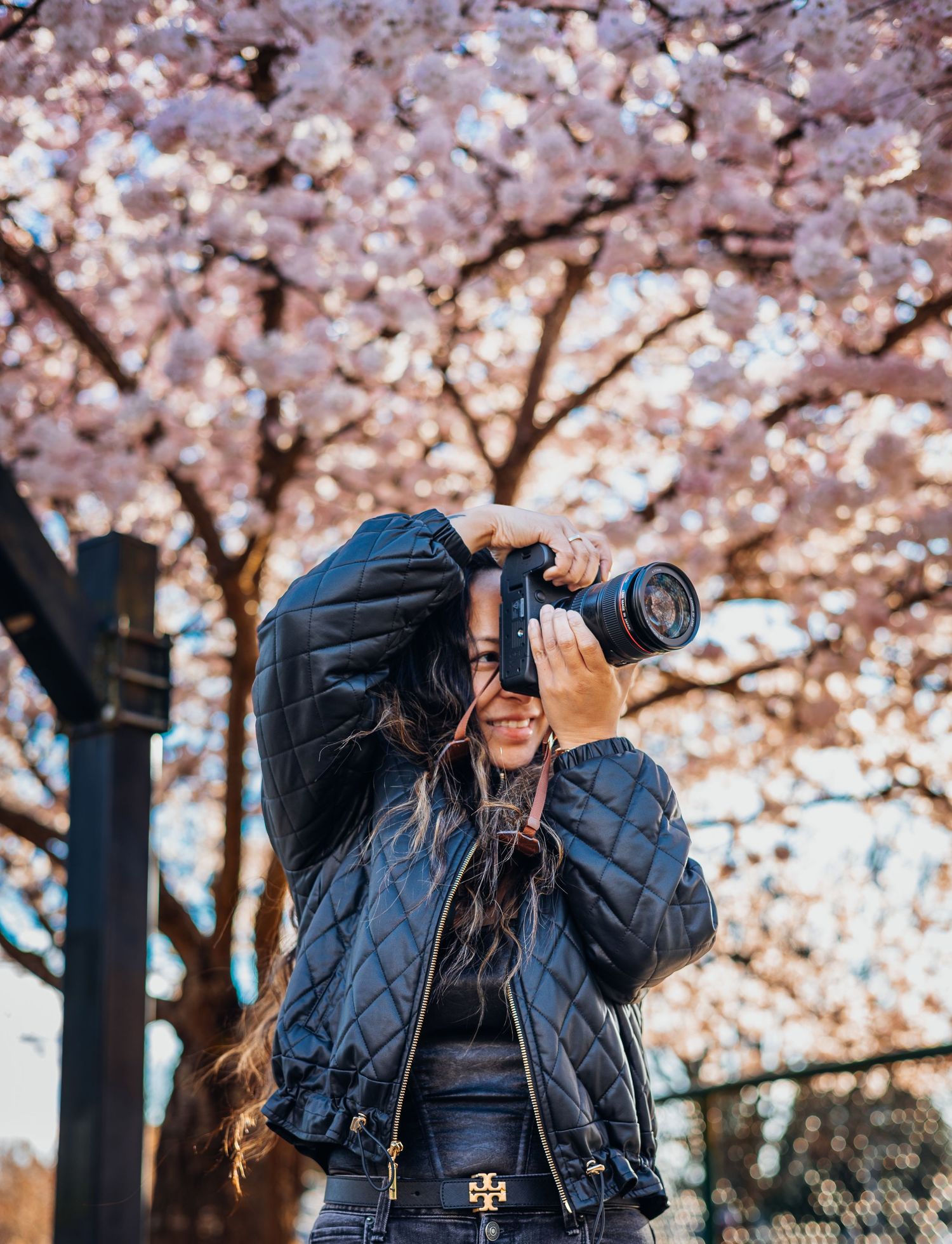 Woman in quilted jacket photographing with DSLR camera under pink cherry blossom trees in spring.
