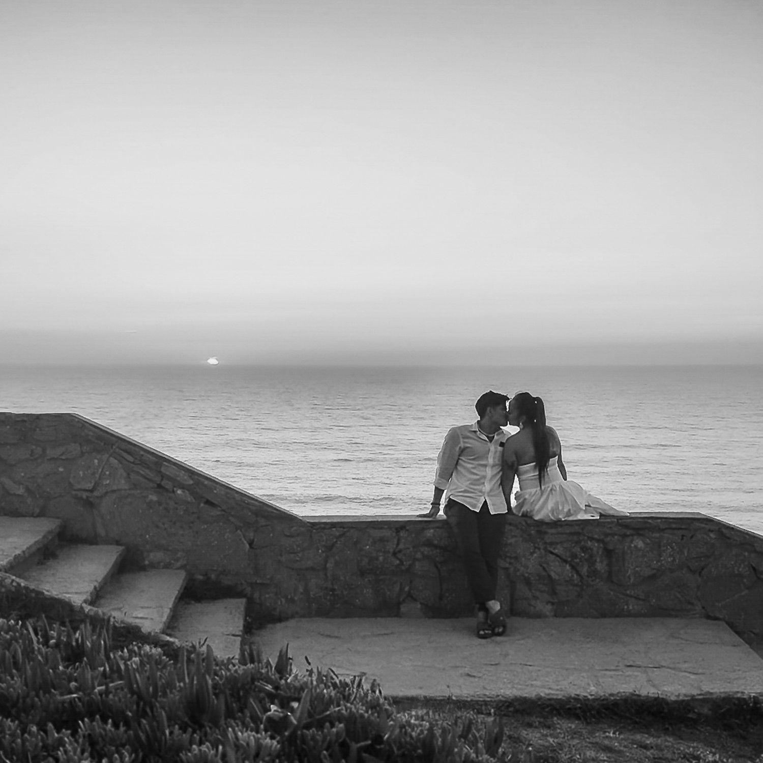 Black and white photo of a couple kissing on a stone wall overlooking the ocean at sunset.
