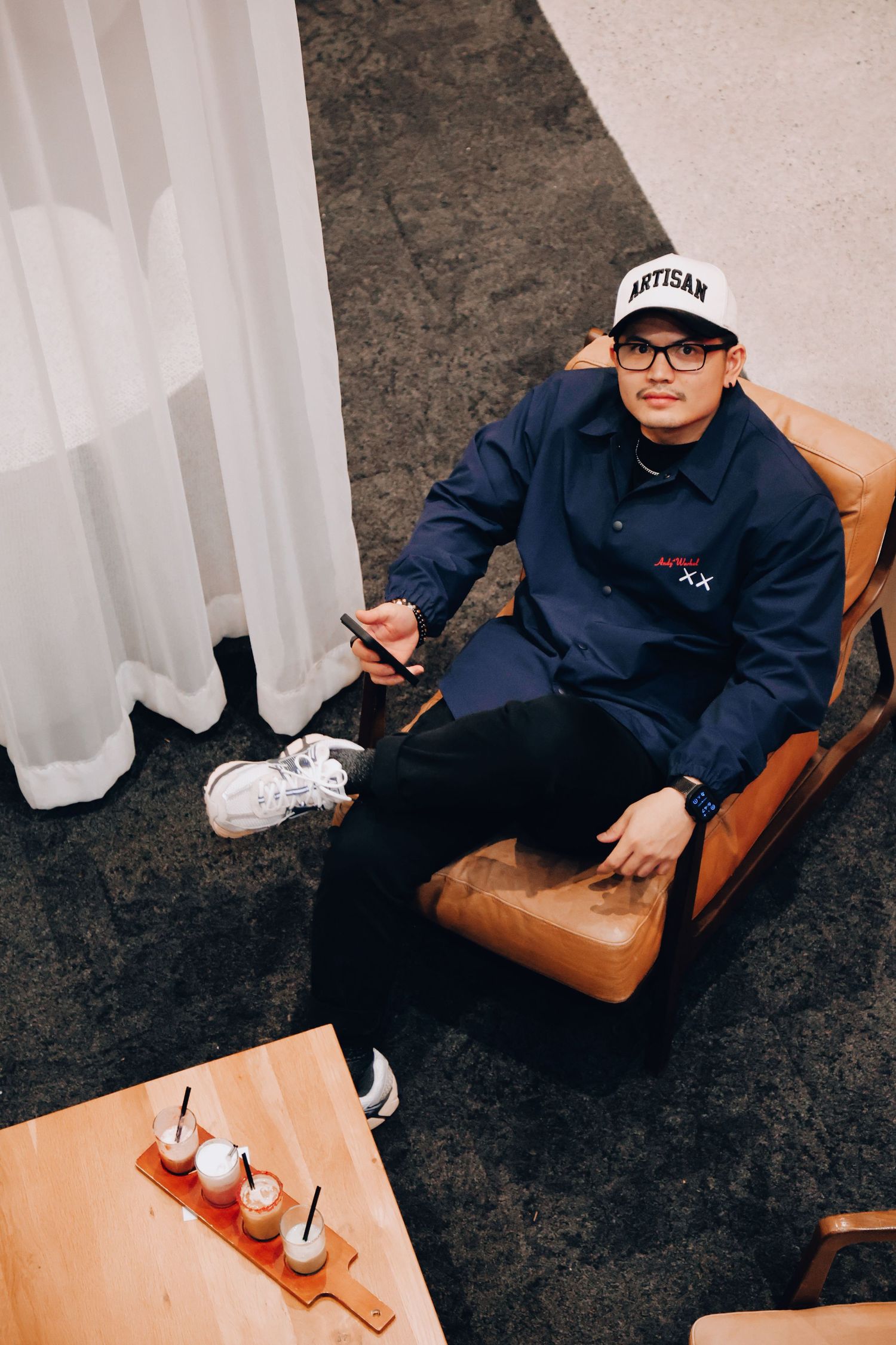 A man in navy Artisan jacket and white cap sits relaxed on leather chair with drinks on table nearby.