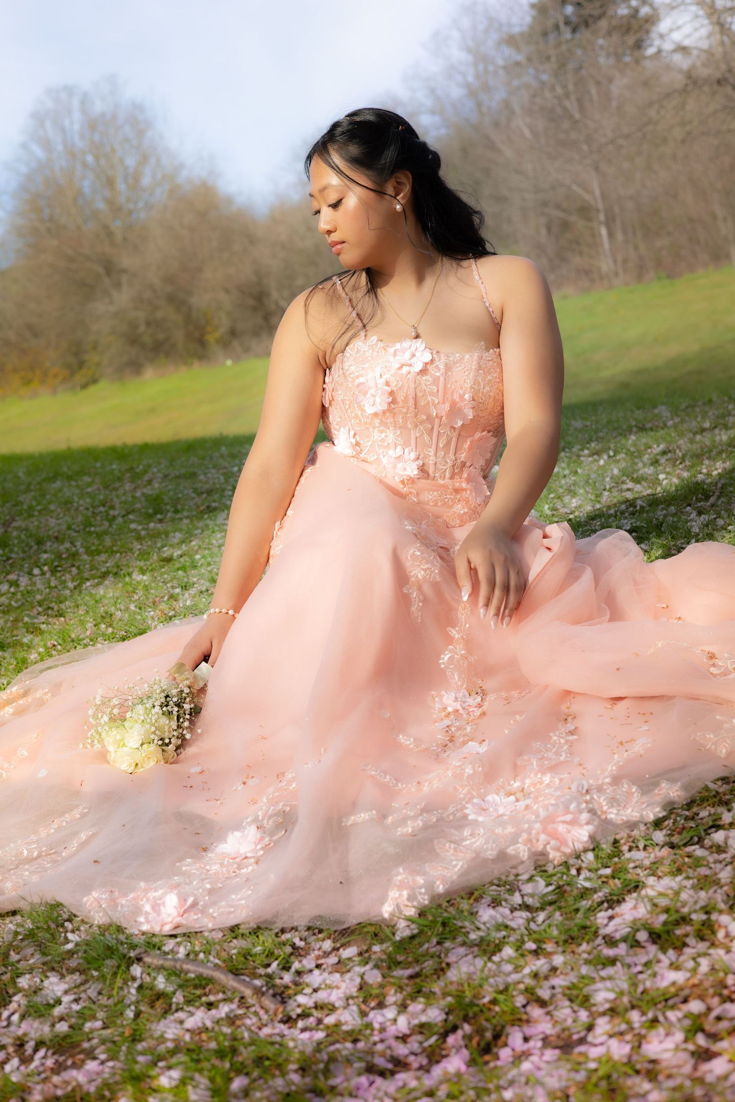 Young woman in a blush pink floral gown sits gracefully on a green meadow holding a bouquet amid fallen petals.
