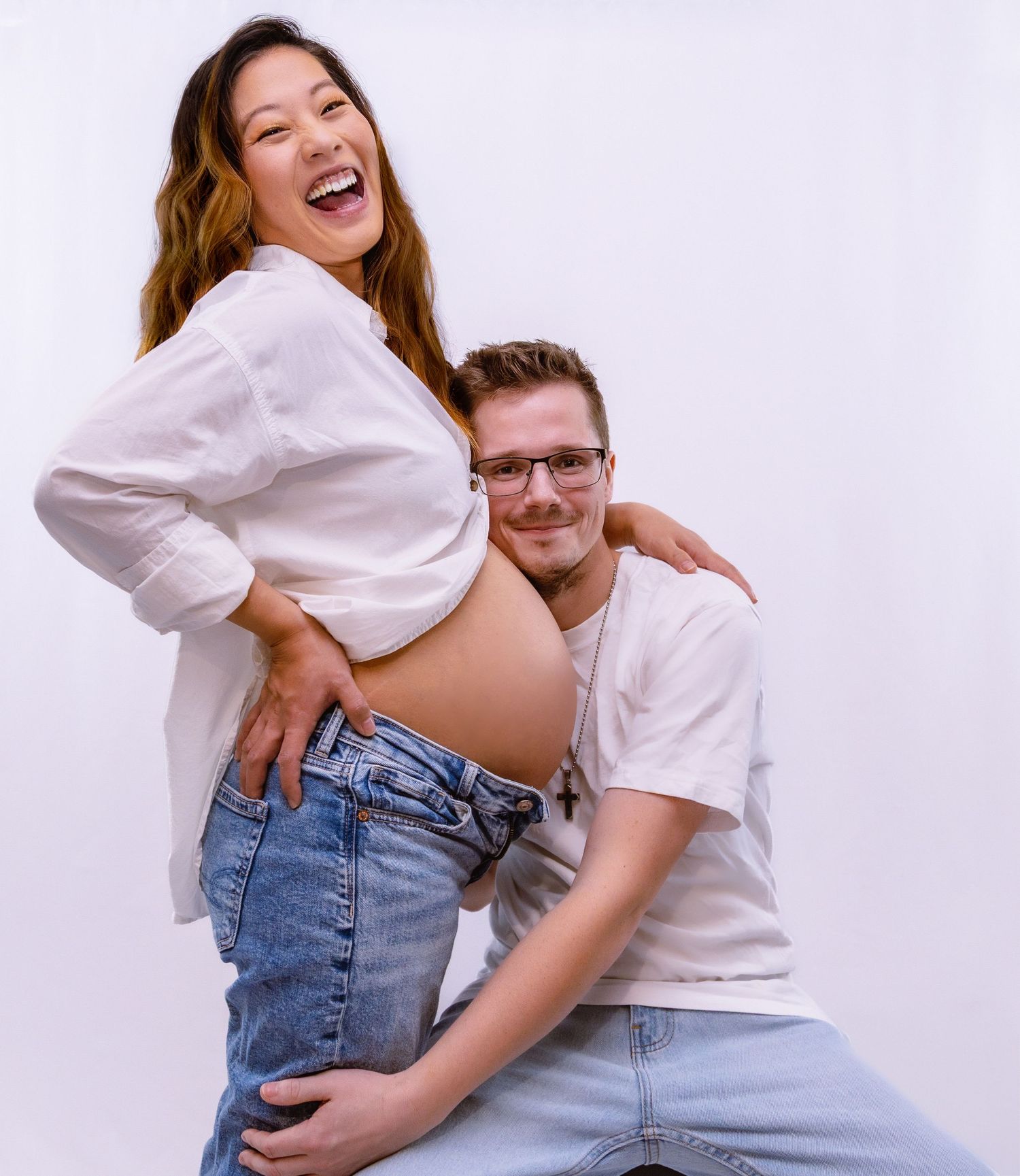 Happy pregnant couple in white outfits laughing together during maternity photo session.