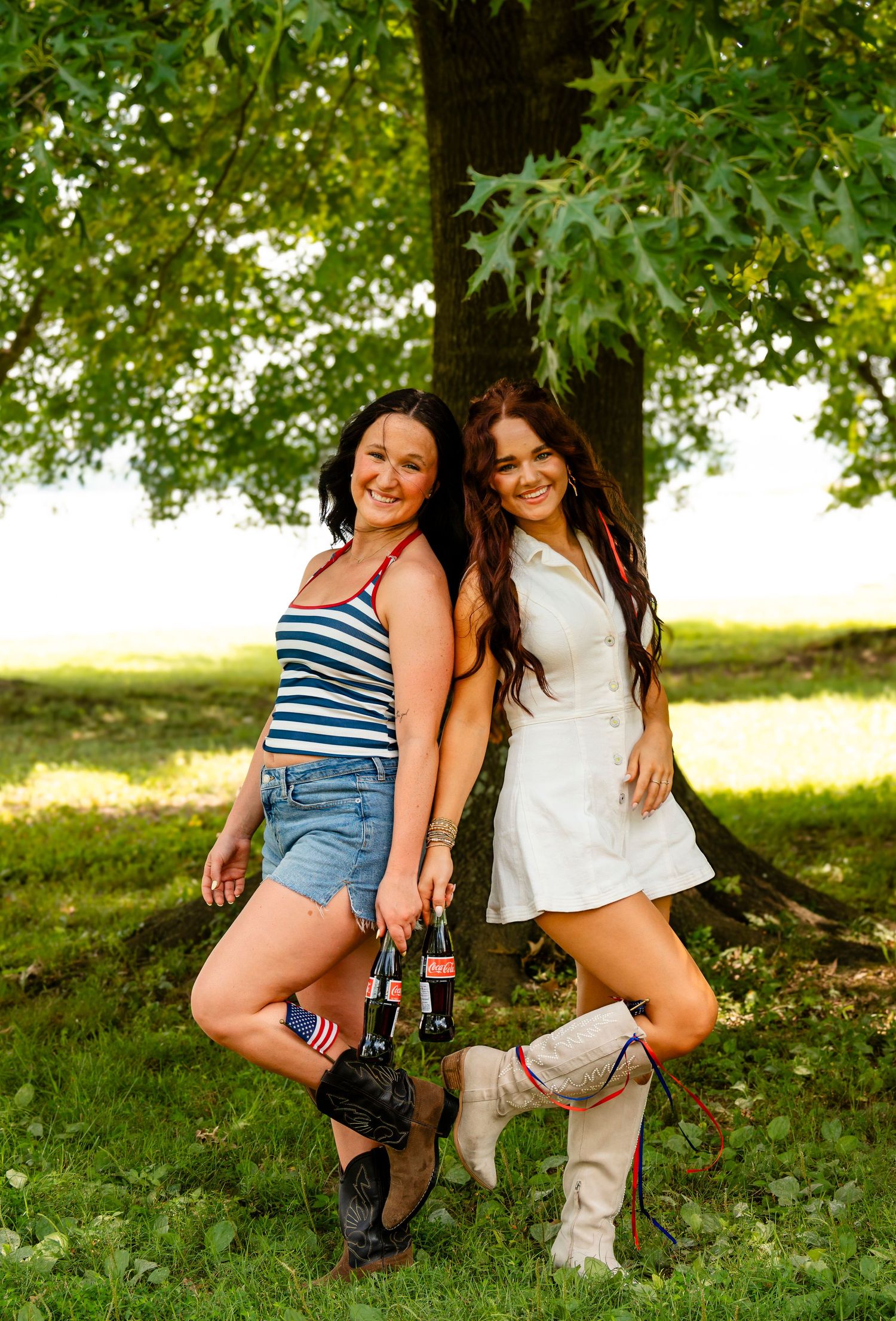 Two women pose by a tree holding Coca-Cola bottles, wearing denim shorts and cowboy boots outdoors.