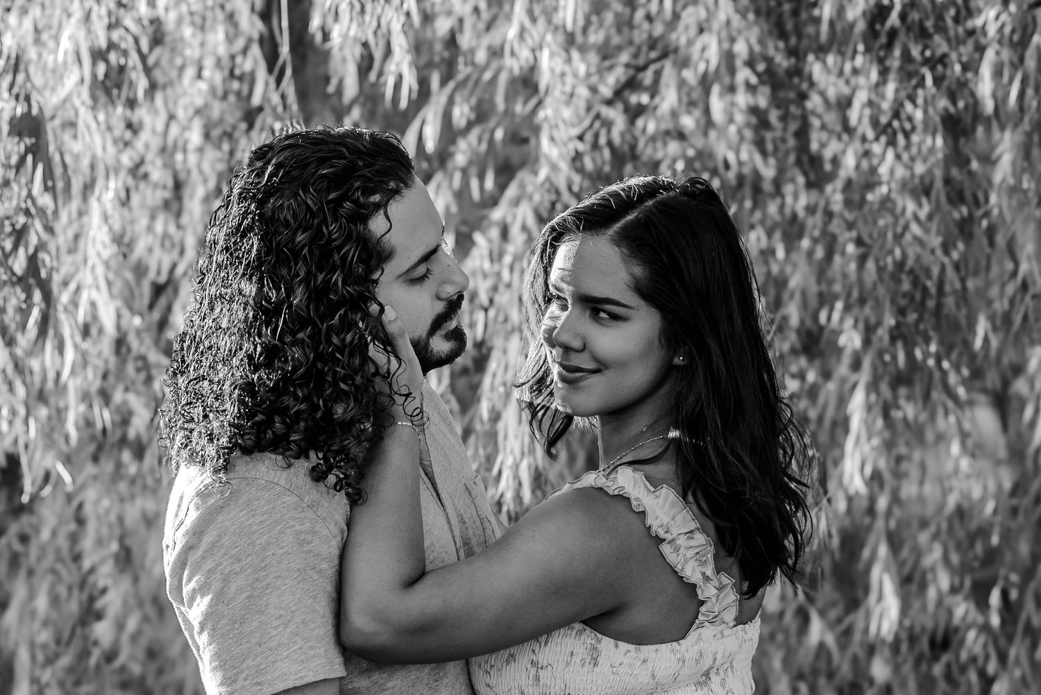 Black and white photo of a romantic couple embracing outdoors under weeping willow tree branches.