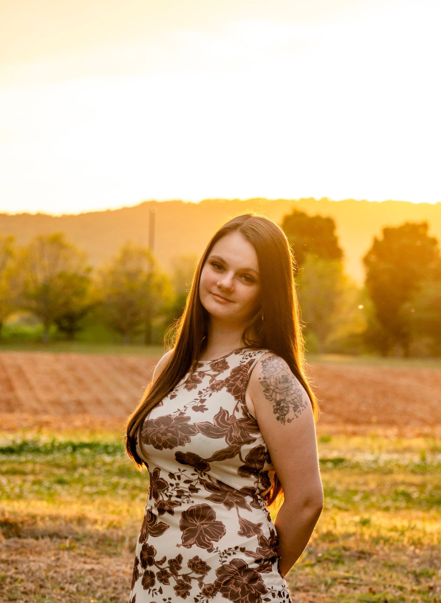 Young woman in floral dress poses at golden sunset in rural field with rolling hills and trees in background.