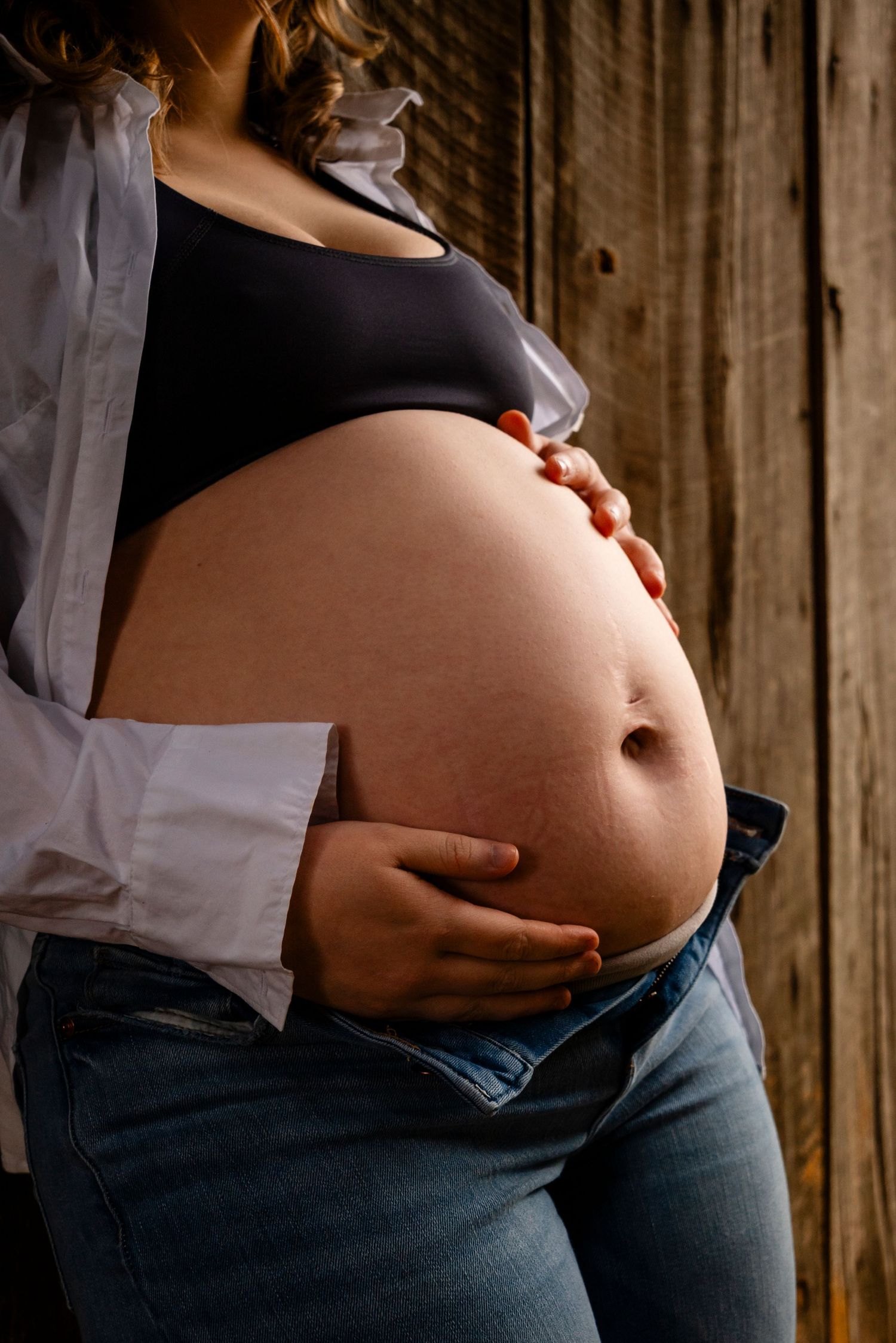 Close-up of pregnant woman in open white shirt and jeans cradling bare baby bump against rustic wooden background.