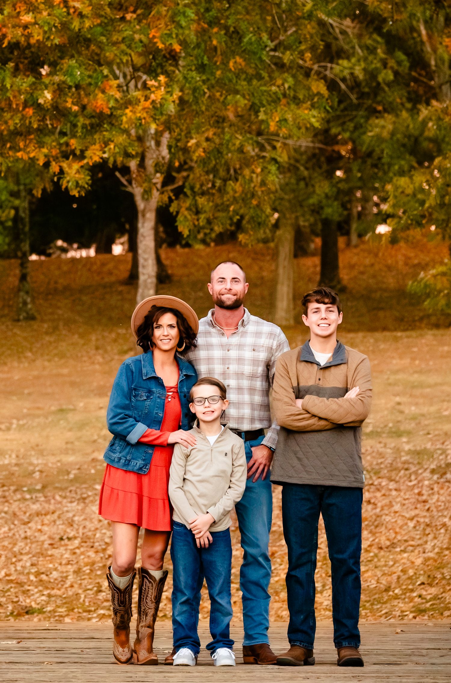 Family of four poses outdoors during fall foliage season on a wooden deck surrounded by colorful autumn trees.