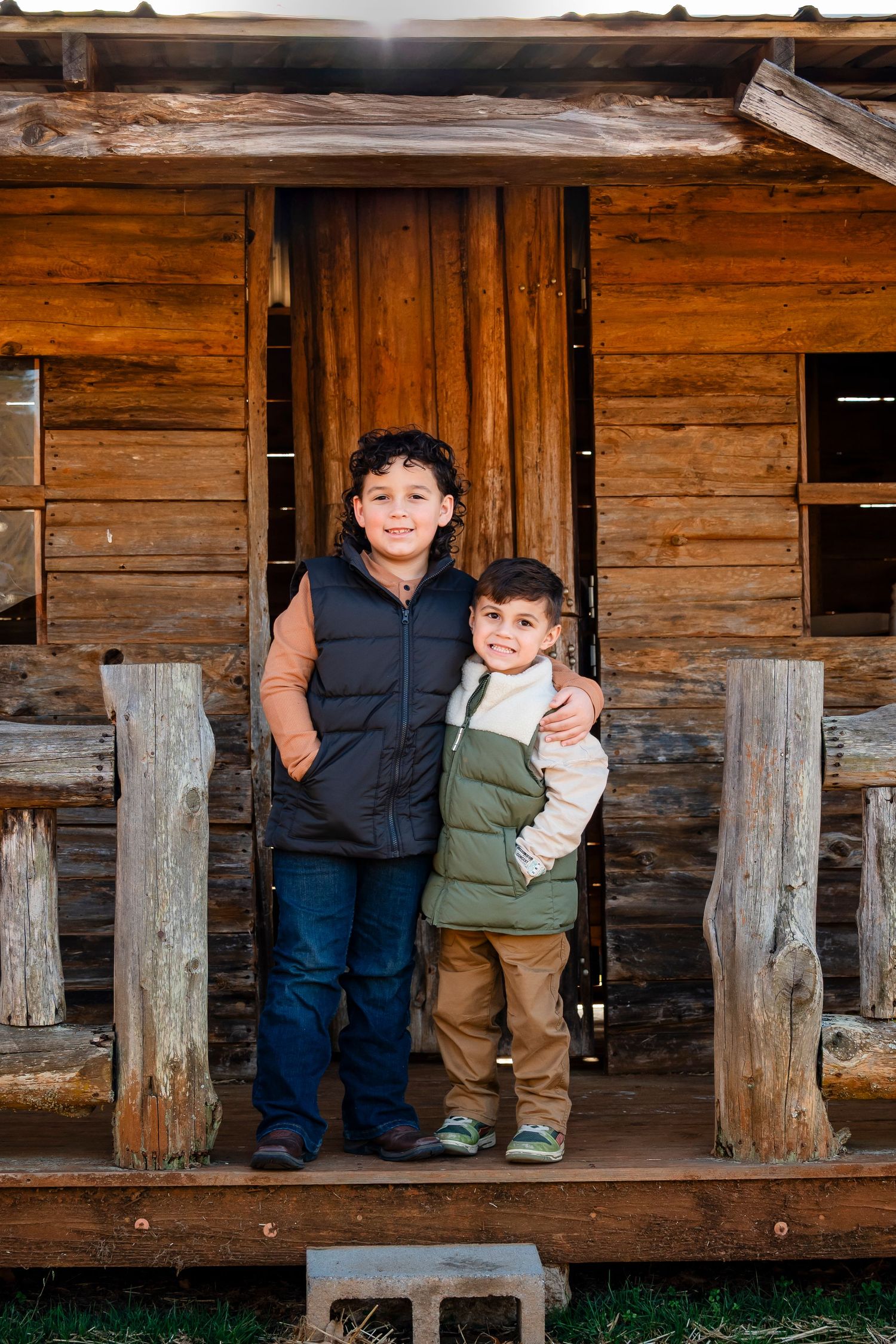 Two young brothers smile together on the porch of a rustic log cabin during a fall family photo session.