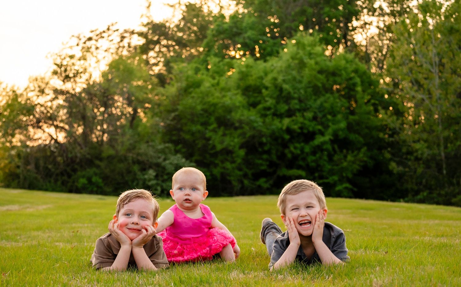 Three siblings lie on green grass smiling, with baby girl in pink dress in the center.