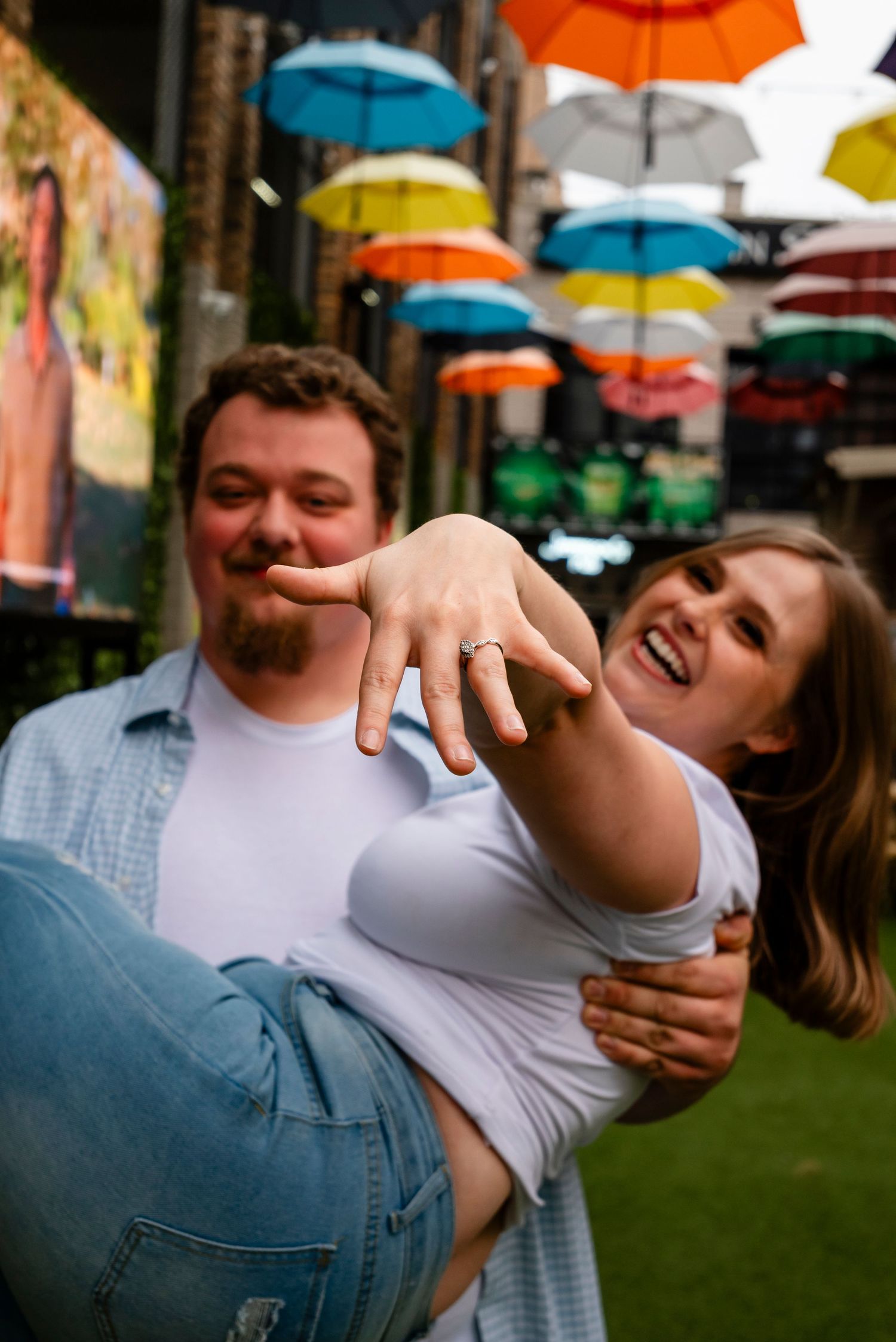 A joyful couple celebrates their engagement under colorful umbrellas, woman showing off her ring while being carried.