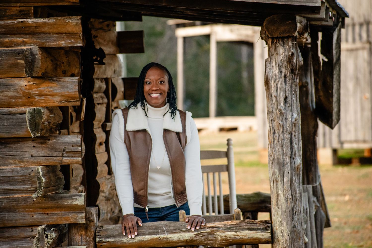 Smiling woman leaning on rustic log cabin porch railing in an outdoor heritage setting.