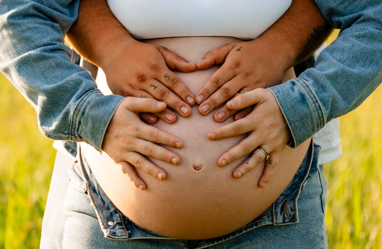 Pregnant woman in denim jacket with two pairs of hands cradling her bare baby bump outdoors in a sunny field.