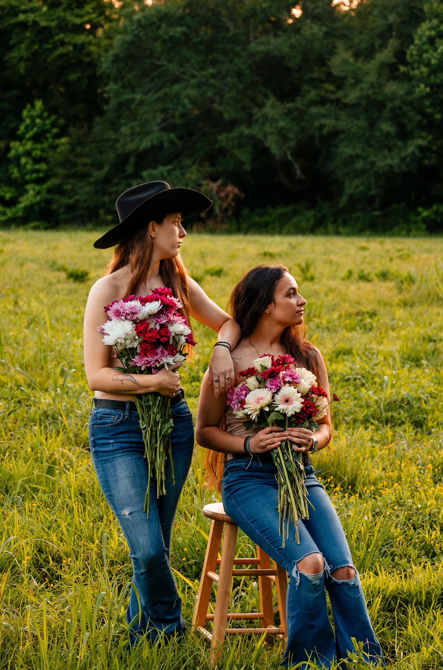 Two women holding colorful flower bouquets pose in a green field, one wearing a black cowboy hat and jeans.