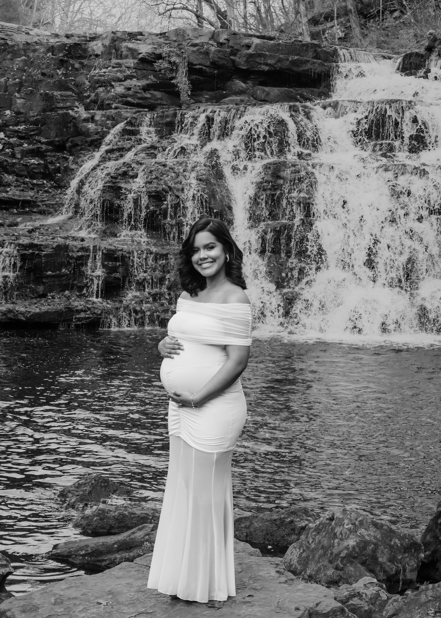 Maternity session with a woman in white off-shoulder gown smiling in black and white photo before waterfall.