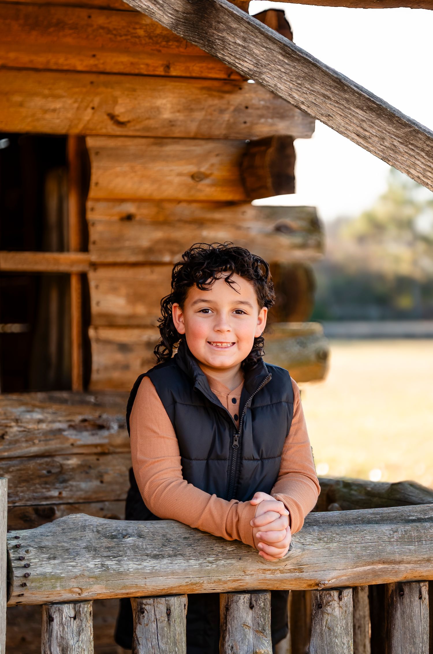 Smiling curly-haired boy leans on a wooden fence rail in front of a rustic log cabin at golden hour.